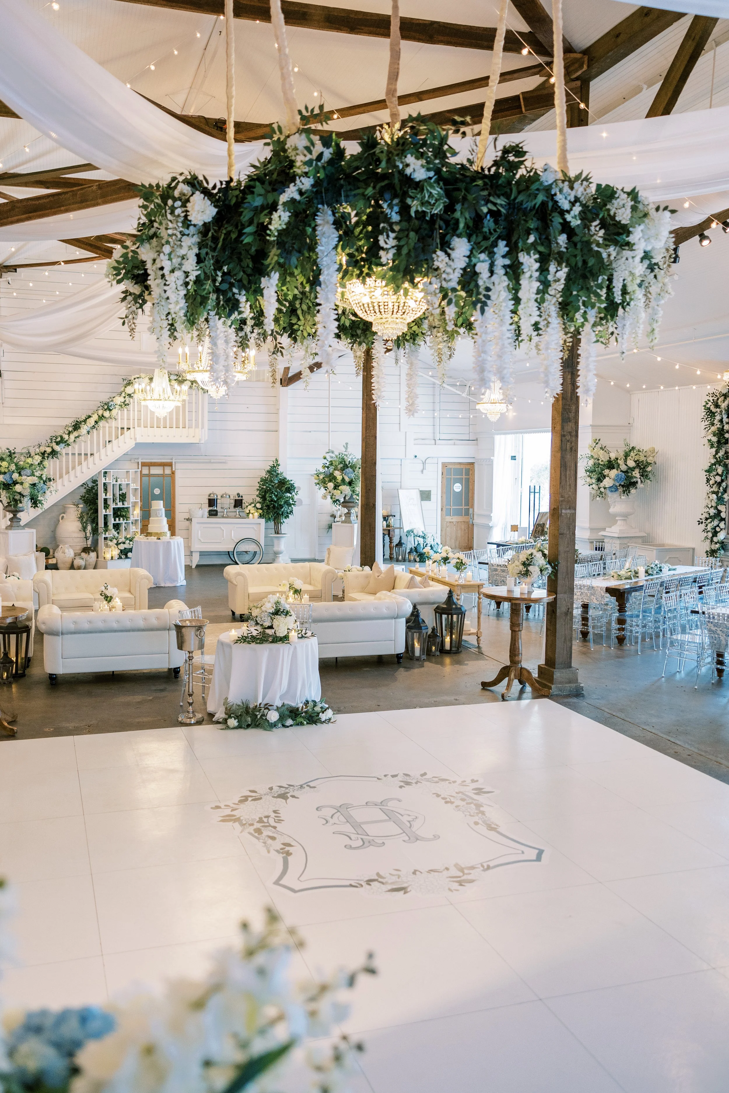 An elegantly decorated wedding reception hall featuring white sofas, floral arrangements, lanterns, and a floral chandelier hanging from the ceiling. There's a center dance floor with a monogrammed design, and the room has draped white fabric and str