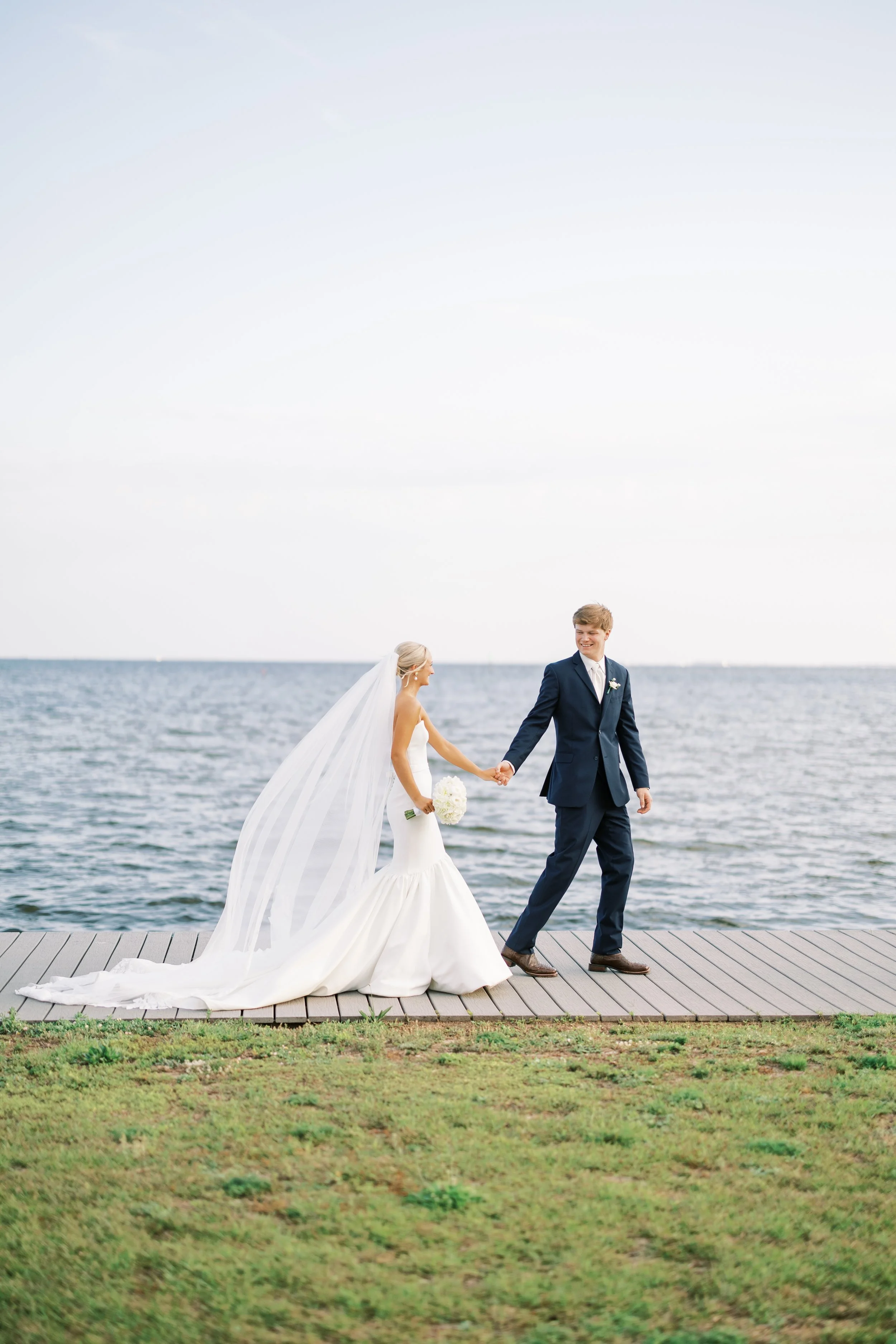 A bride and groom walking hand in hand along a dock by the water, with the bride in a white wedding dress and veil holding a bouquet, and the groom in a navy suit, during their wedding.