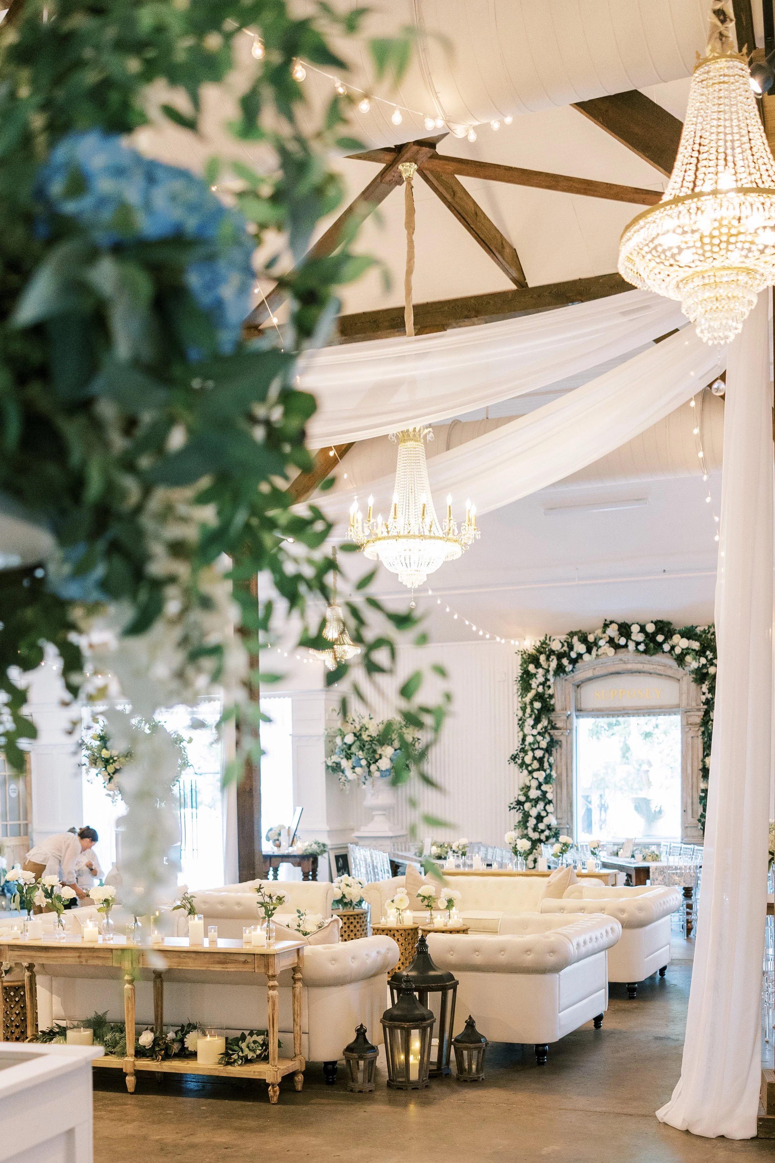 Elegant event space with white sofas, floral arrangements, chandeliers, candles, and draped fabric, decorated for a celebration.