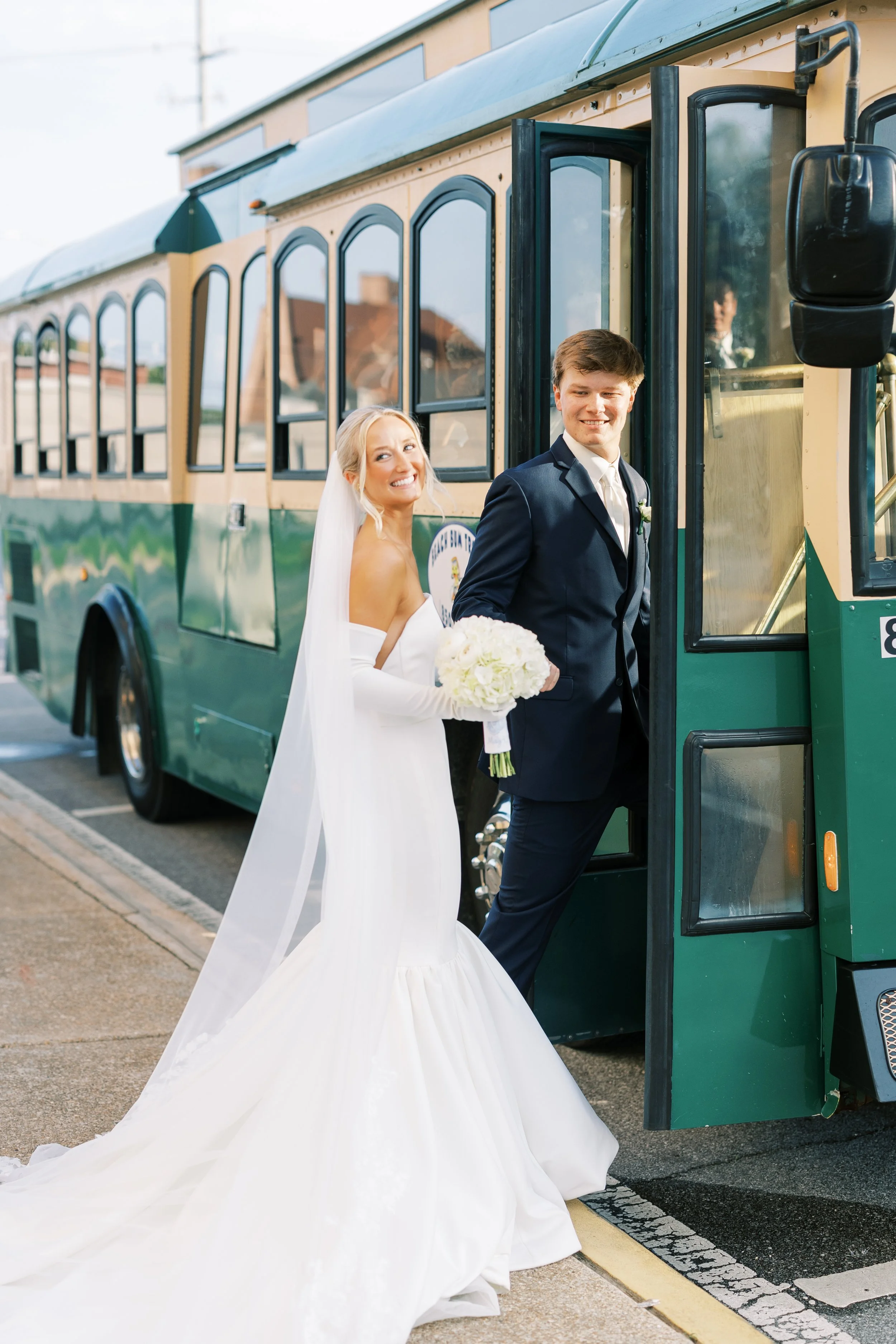 A bride and groom step off a green trolley bus on their wedding day. The bride is in a white wedding gown holding a bouquet, and the groom is in a navy suit.