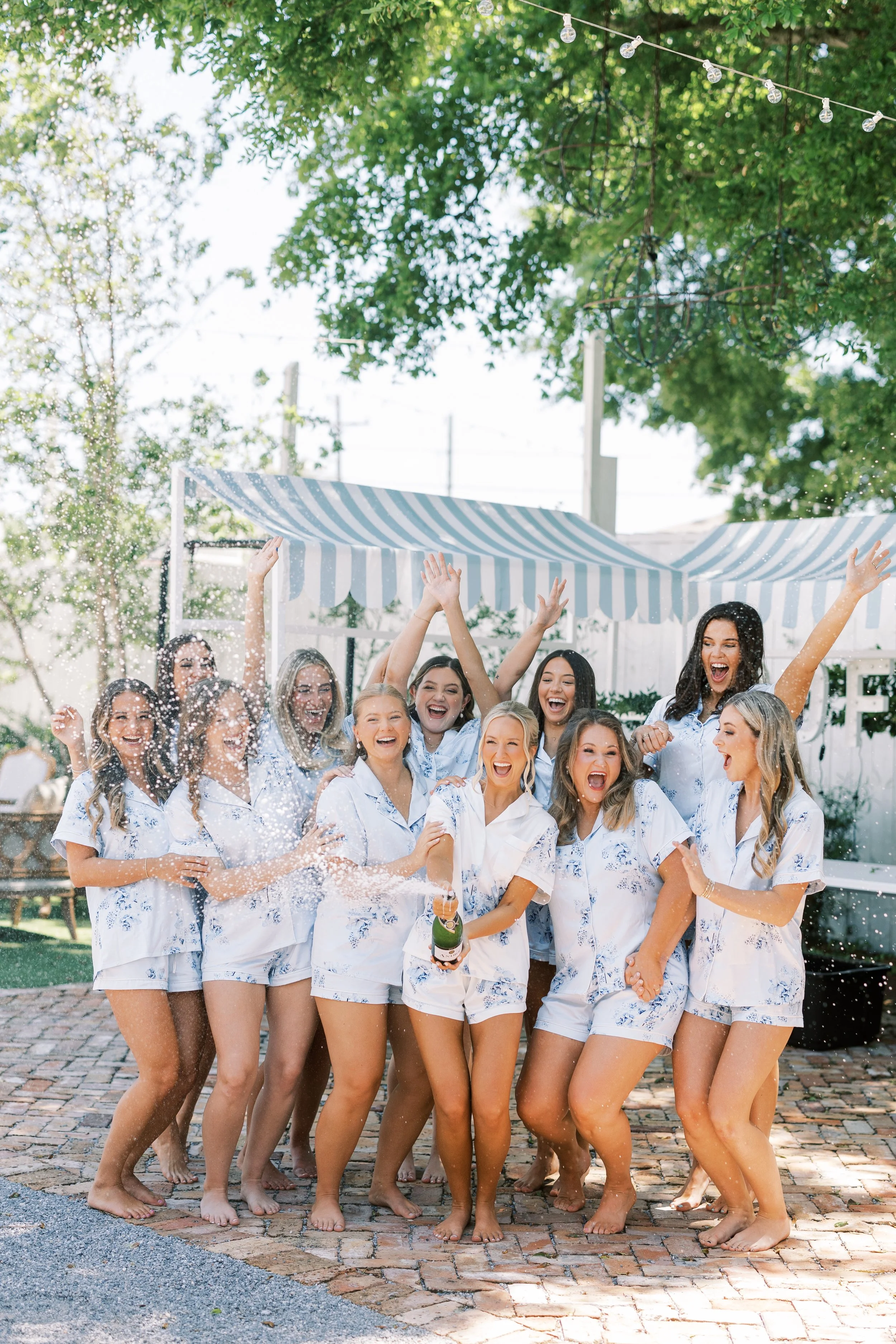 A group of women celebrating outdoors, dressed in matching pajamas, with some holding a bottle of champagne and others throwing water or champagne, under string lights and a striped canopy.