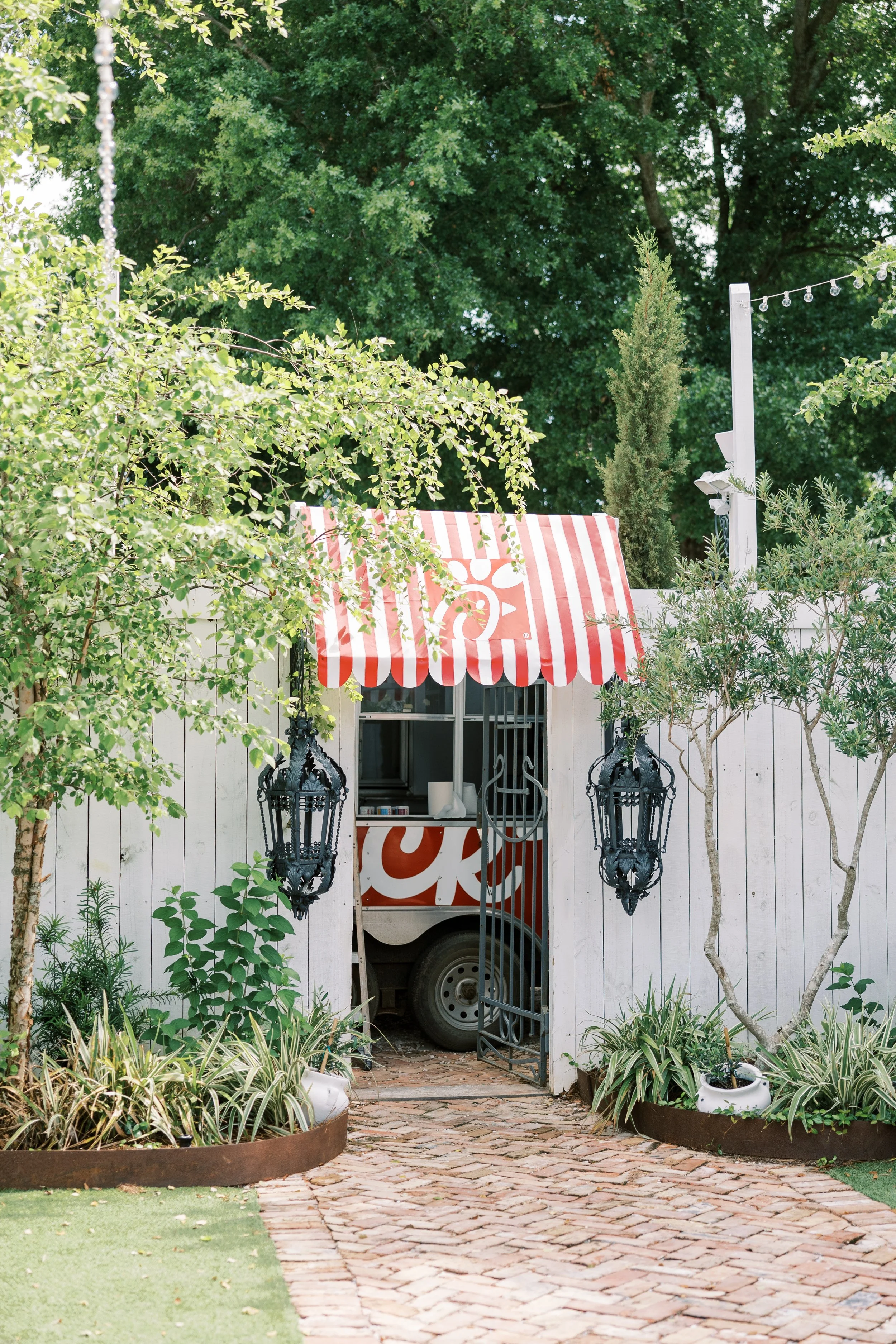 A small food or ice cream truck with a red and white striped awning is partially hidden behind a white fence and surrounded by lush green trees and plants. There are two black ornate lanterns hanging on either side of the gate, and a brick pathway le