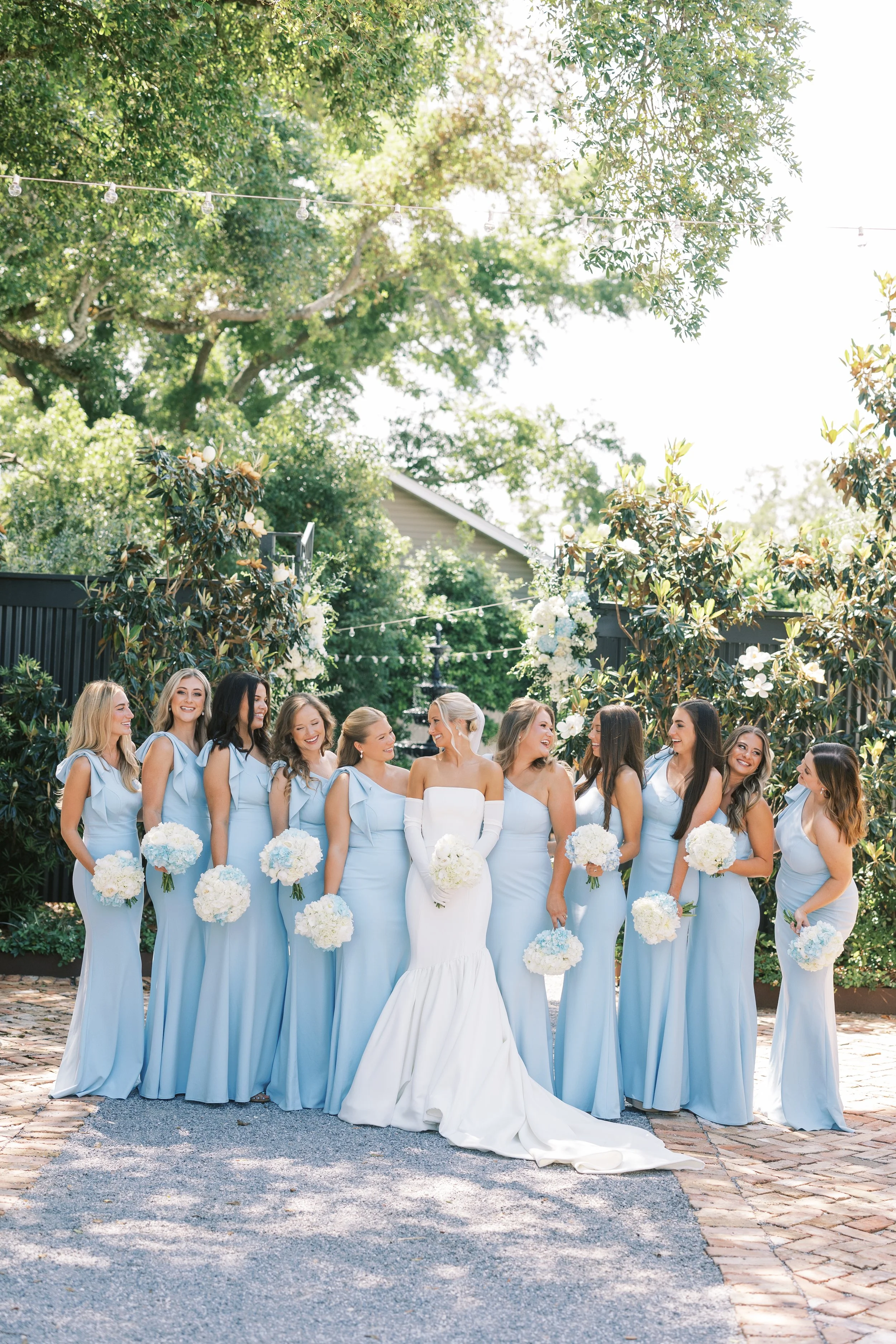 Bride and bridesmaids outdoors, a bride in a white gown, seated middle, surrounded by bridesmaids in matching light blue dresses, holding white bouquets, under green trees, sunny day.