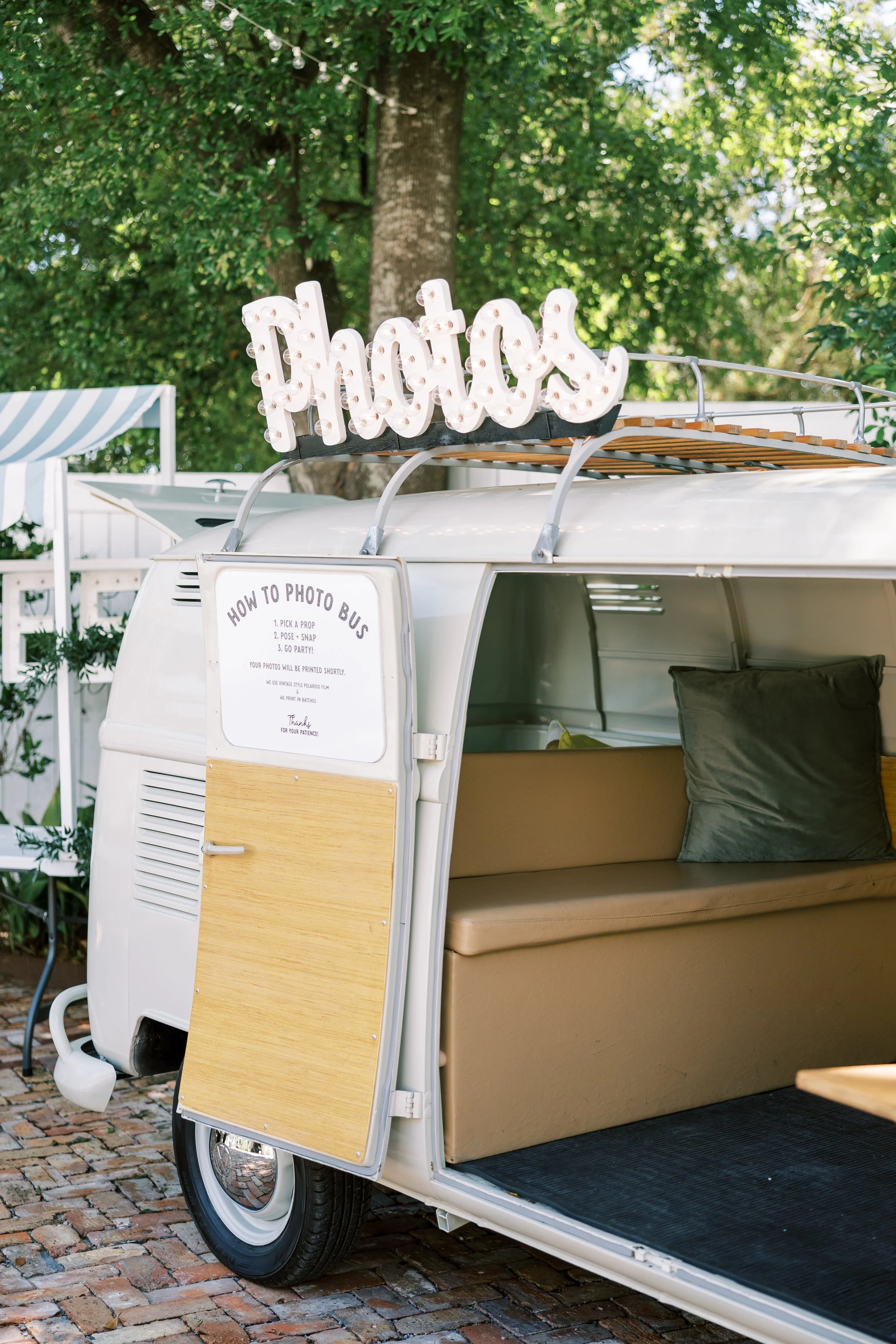 A vintage photo booth on wheels with a illuminated 'Photos' sign on top, situated outdoors with trees and a striped canopy in the background.