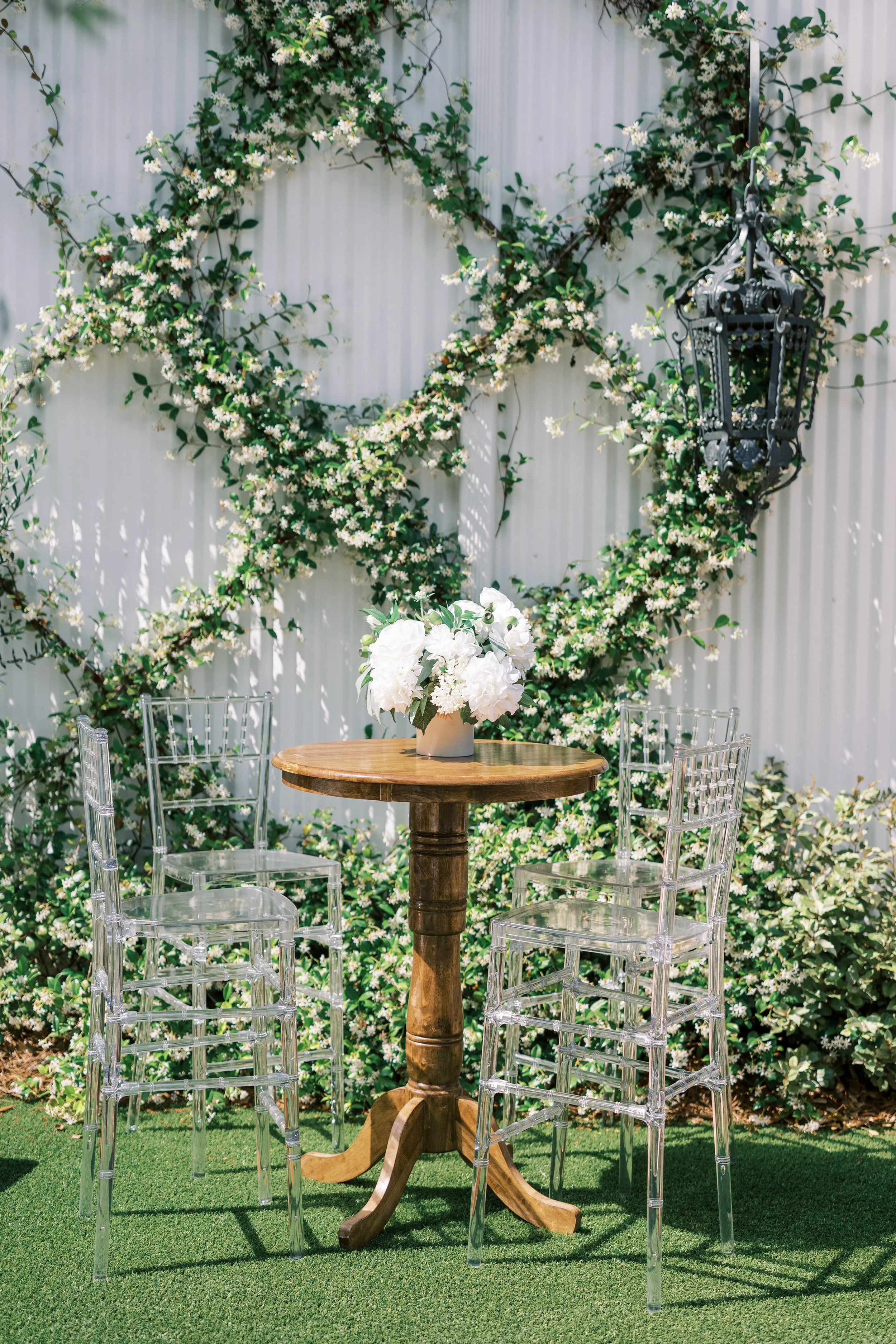 A round wooden table with a white flower arrangement in a vase, flanked by transparent acrylic chairs, set against a backdrop of a white wall with green foliage and white flowers, and a black decorative lantern hanging.