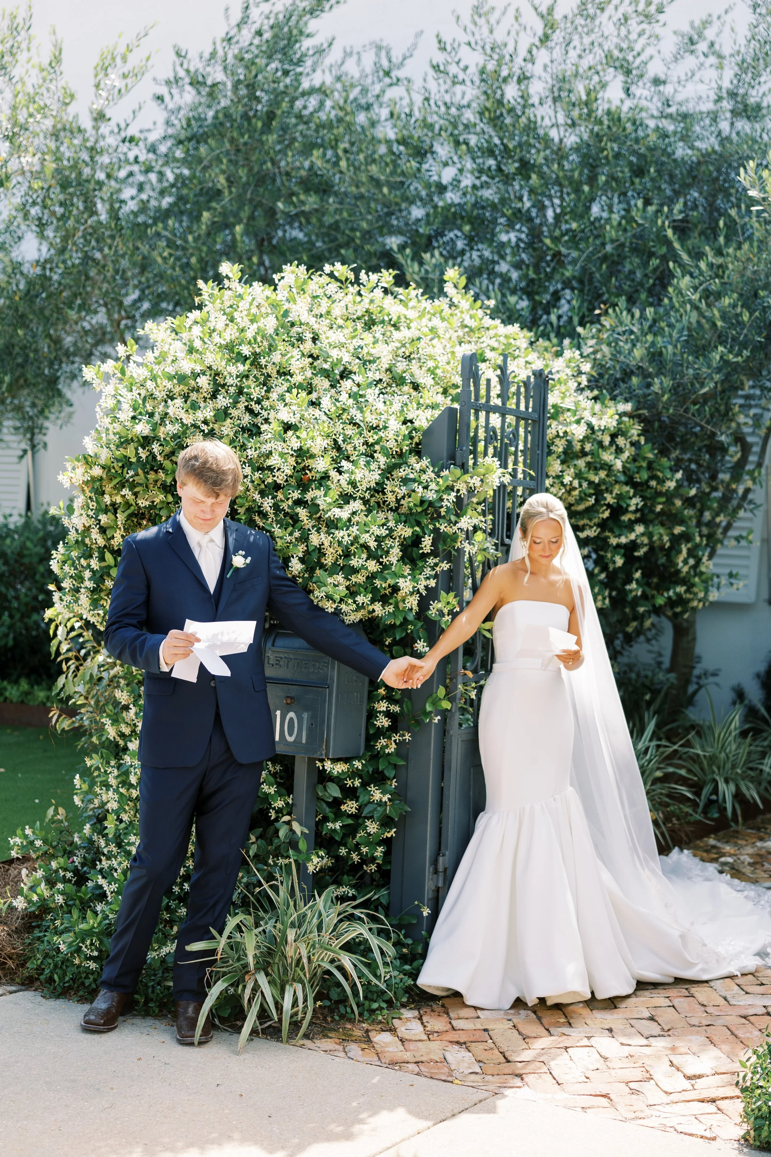 A bride and groom holding hands in front of a lush, blooming hedge, with the groom reading vows and the bride looking at her vows, during their wedding ceremony.