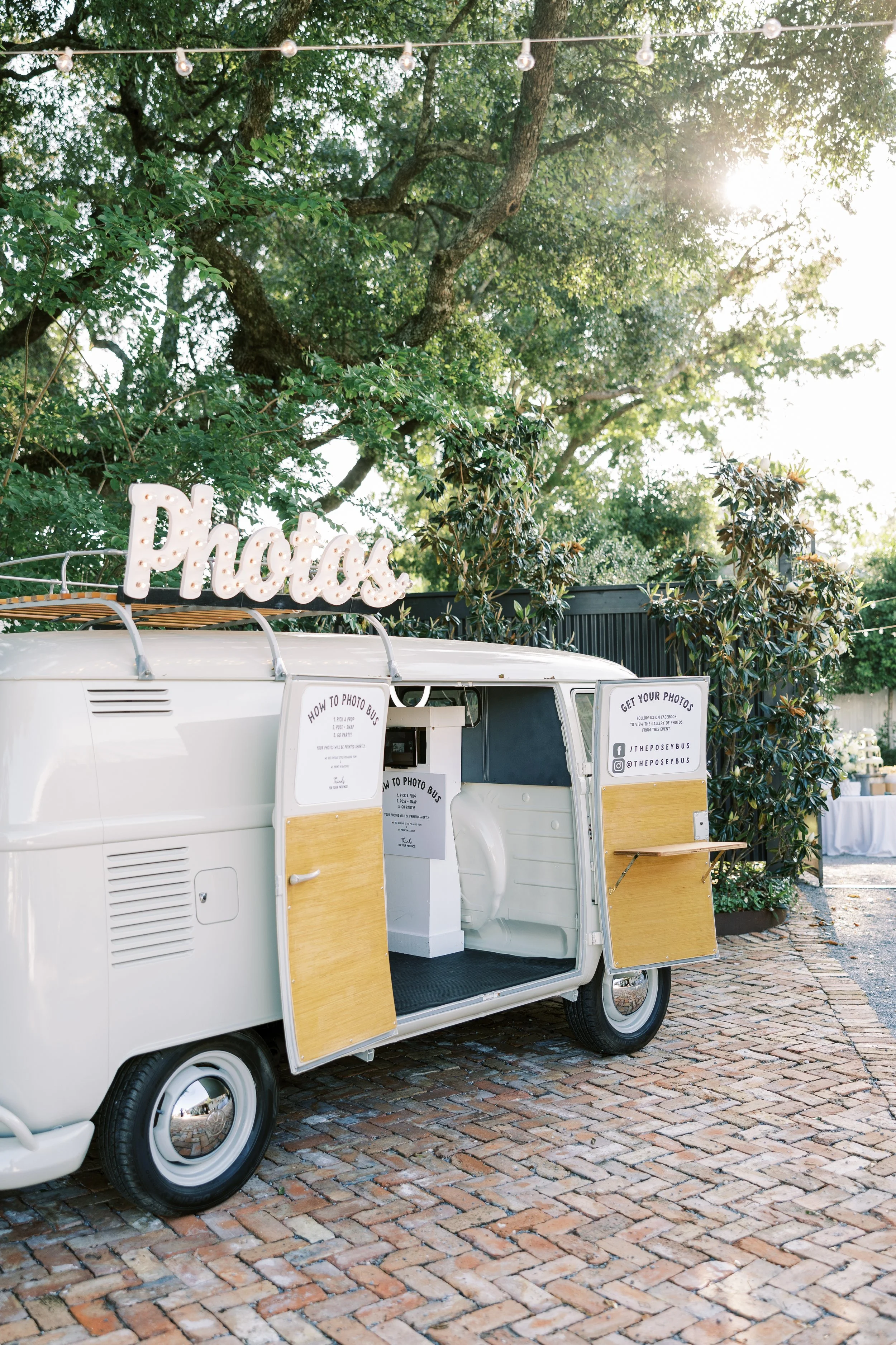 A vintage white van serving as a photo booth at an outdoor event, with a lighted 'Photos' sign on top, open side doors, and signs providing instructions on how to use the photo booth.