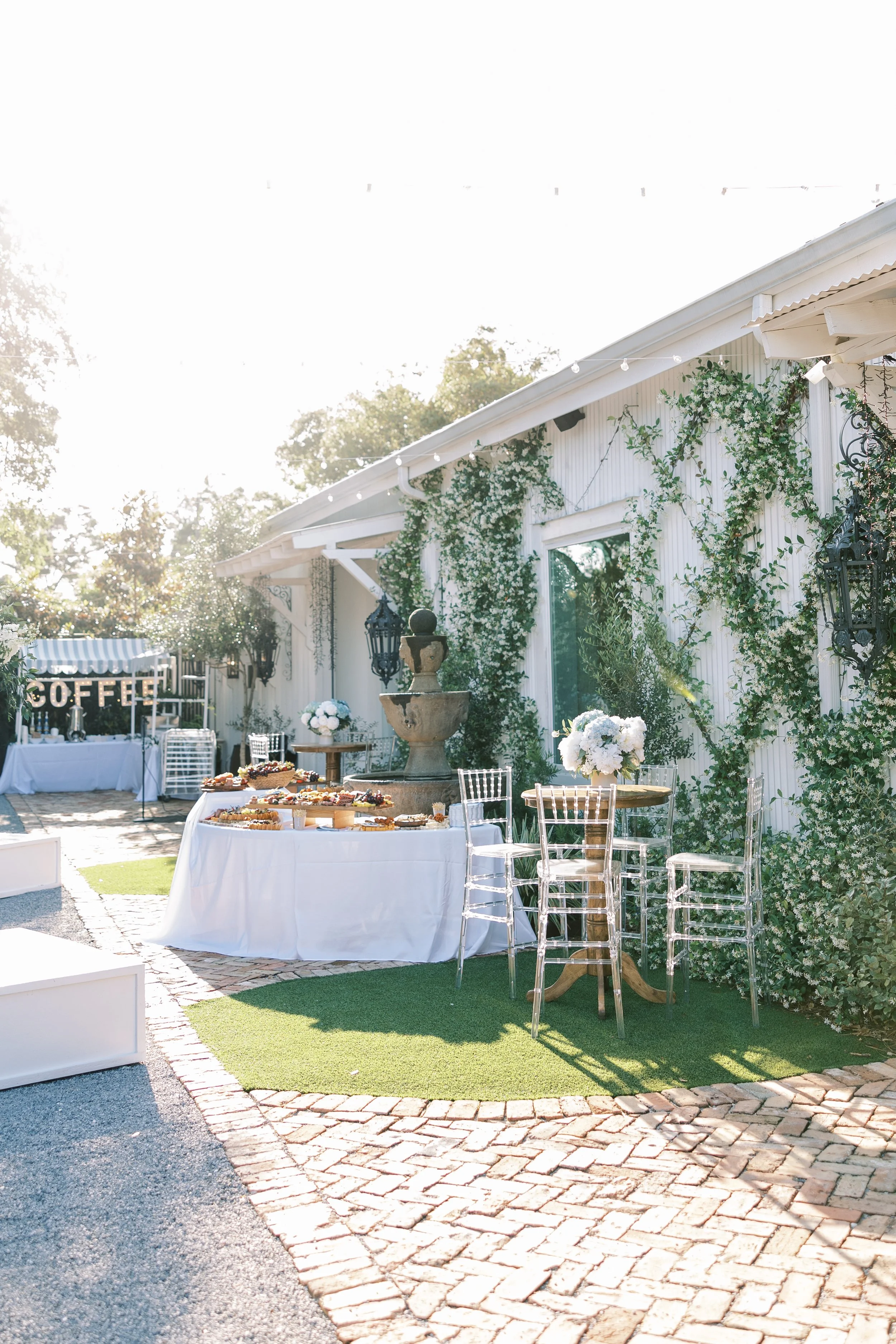 Outdoor Pensacola celebration setup with tables, flowers, and a coffee station, decorated with green vines and string lights, on a paved brick patio.