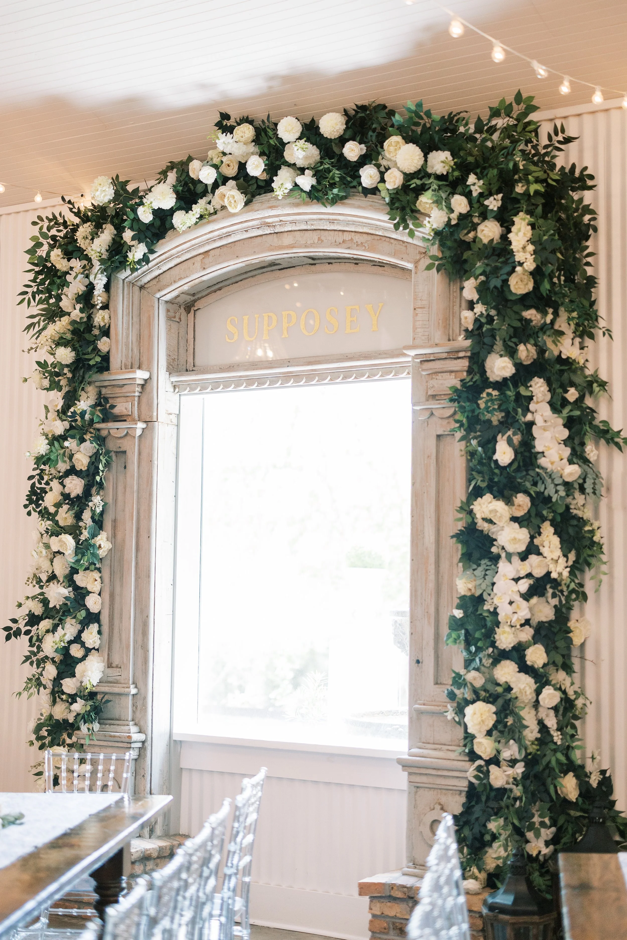 Decorated archway with white flowers and greenery, with the name 'SUPPOSEY' on a sign above a window.