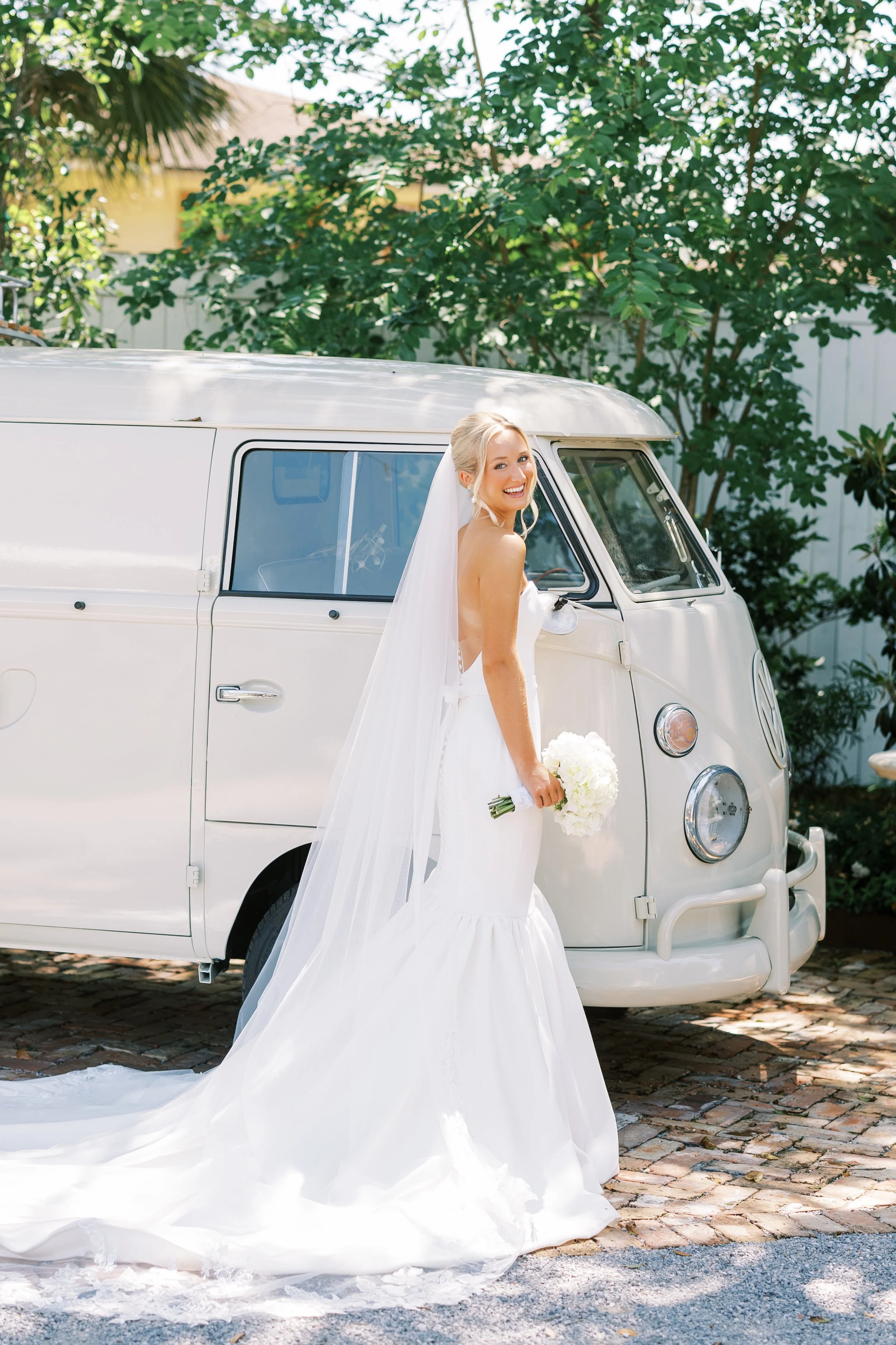 A smiling bride in a white wedding gown holding a bouquet stands outdoors beside a vintage white van on a cobblestone driveway, with green trees in the background.