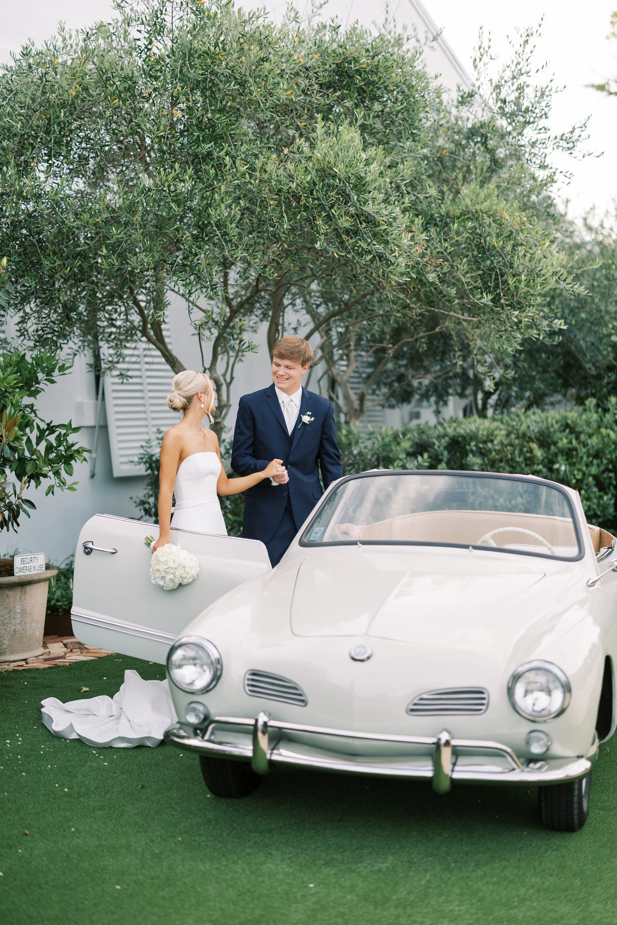 A bride and groom smiling and holding hands next to a vintage white convertible car on a green lawn, with trees and a building in the background of a Pensacola Wedding Venue