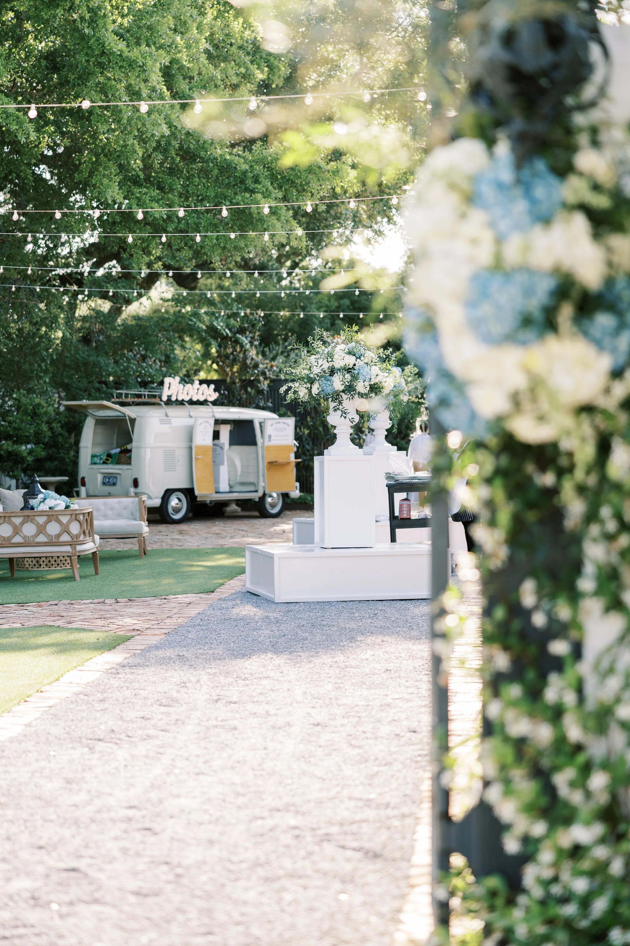 Decorative setup for an outdoor event with white floral arrangements, a vintage photo booth, and string lights hanging overhead in a Pensacola Wedding Venue.