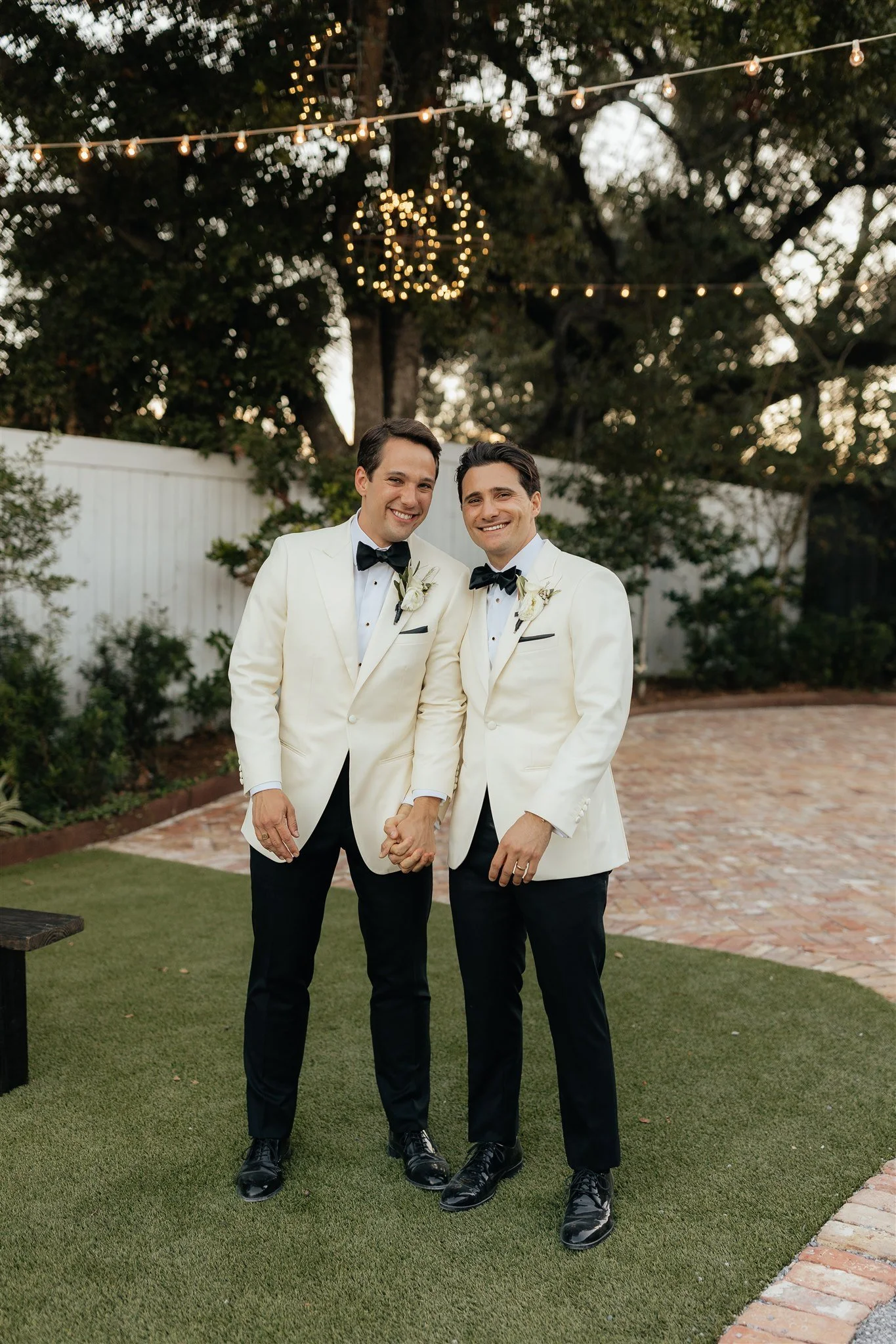 Two men wearing white tuxedos with black bow ties and black pants standing outdoors, holding hands and smiling, with decorative string lights and a wooden fence in the background.