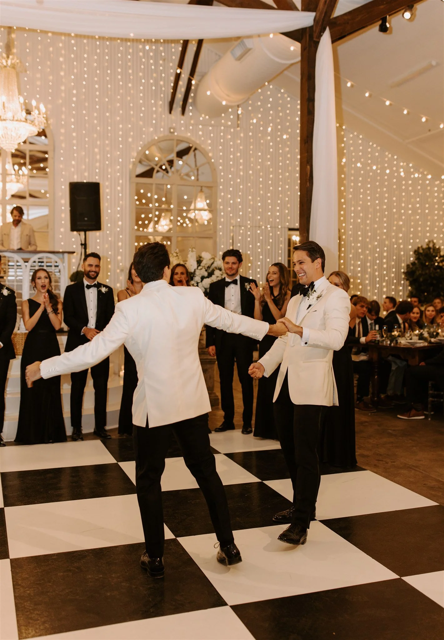 Two men in tuxedos dancing and holding hands at a wedding reception with guests in the background, decorated with string lights and chandeliers.