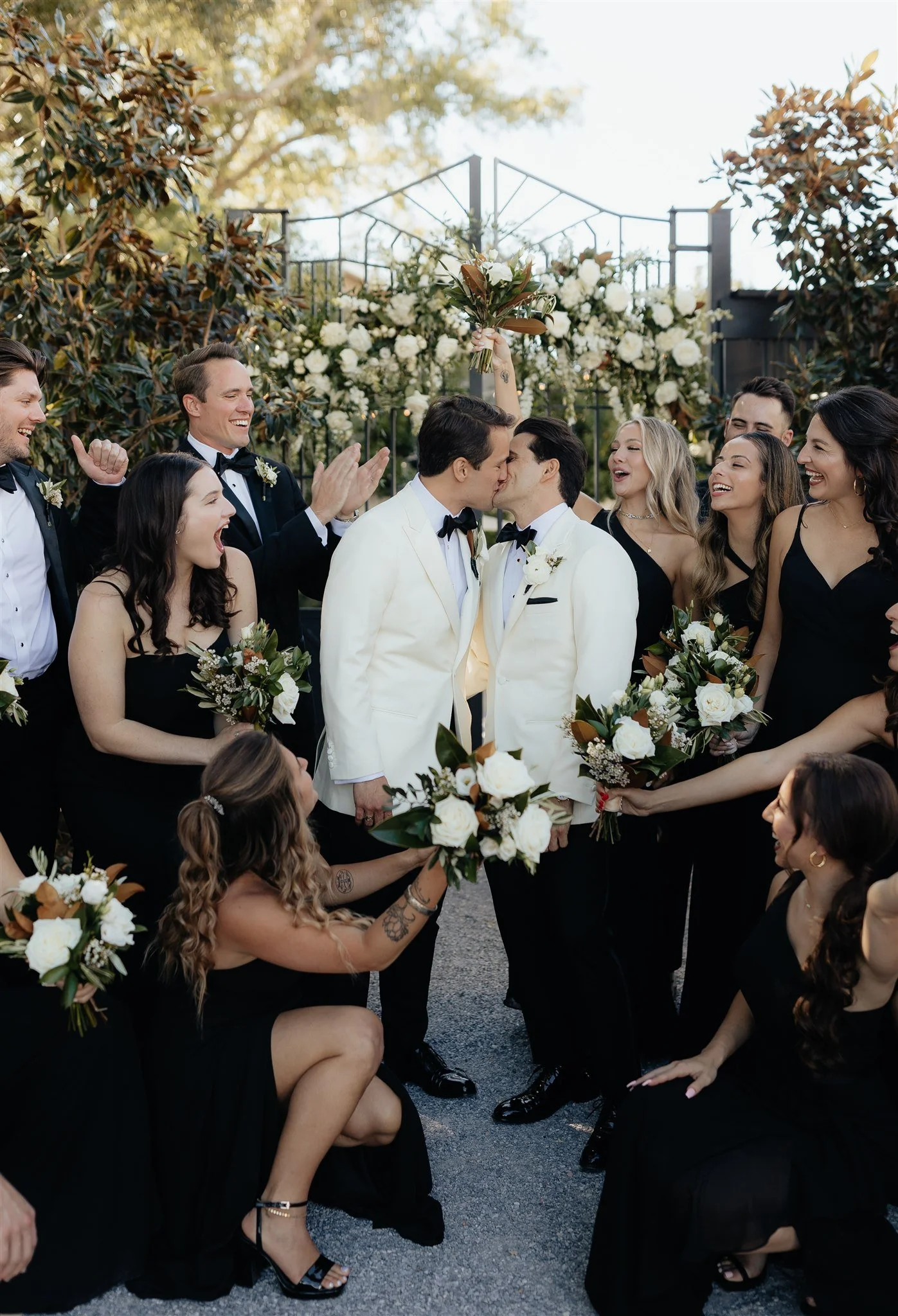 Two men in white tuxedos kiss in the center of wedding party holding bouquets, surrounded by smiling bridesmaids and groomsmen outdoors on a sunny day.