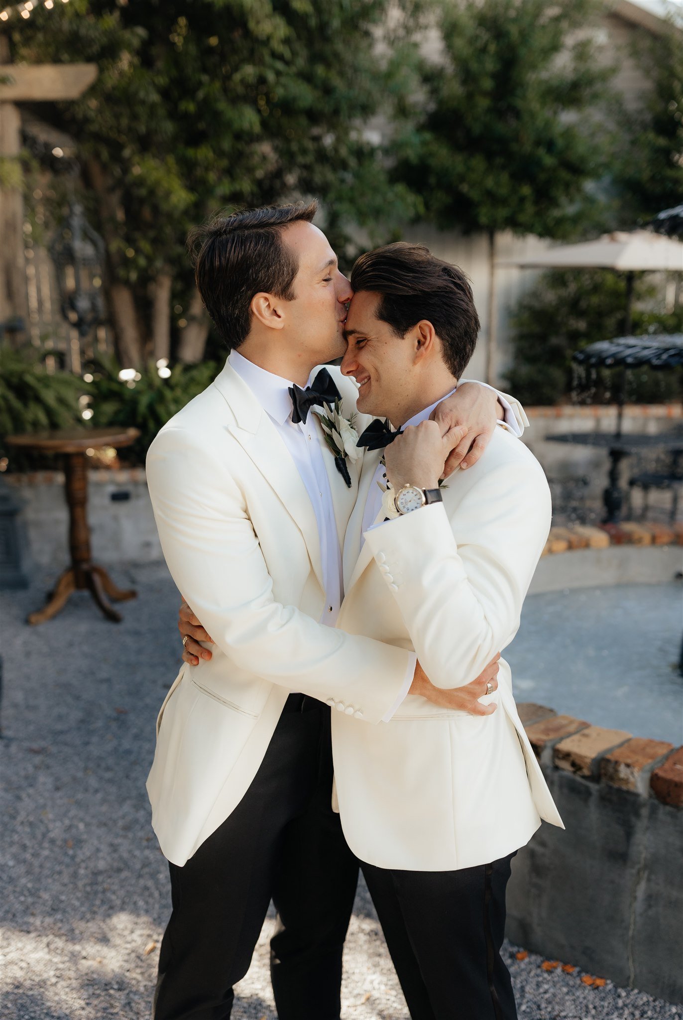 Two men in wedding tuxedos embracing, one kissing the other's forehead, in an outdoor garden setting with trees and a fountain in the background.