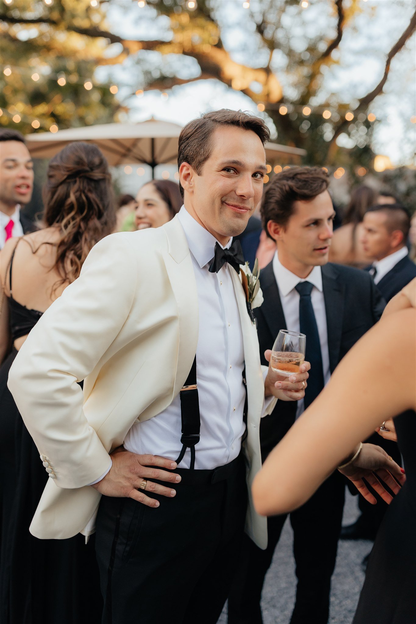 A man in a white tuxedo jacket, black bow tie, and suspenders holding a glass of wine at an outdoor evening event with string lights and other people in formal attire.