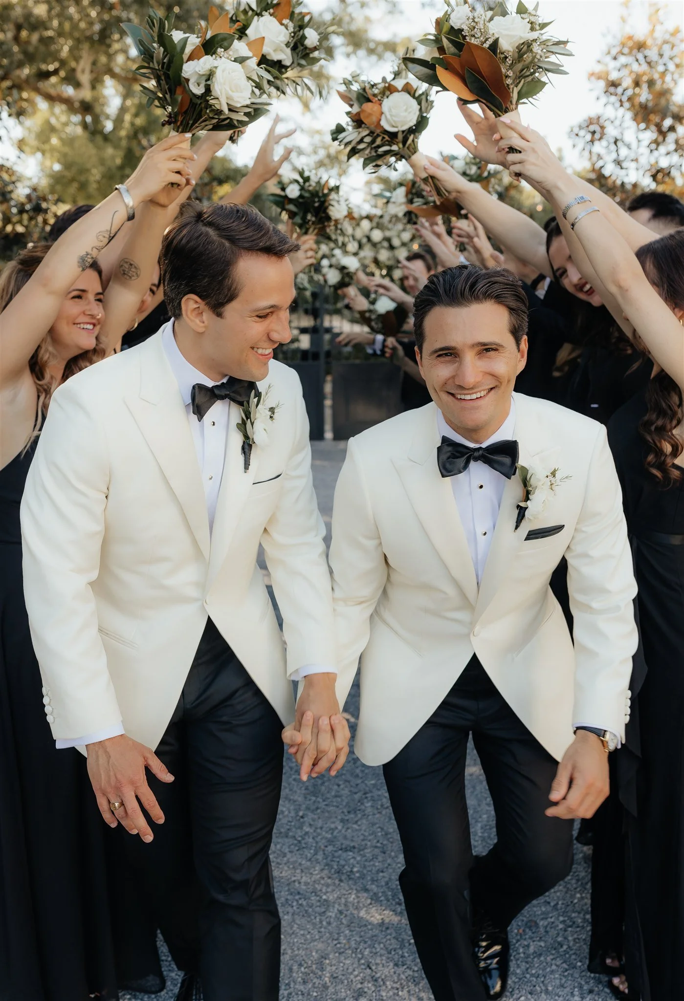 Two men in white tuxedos and black bow ties holding hands, walking through a line of women with raised arms holding bouquets, at a wedding celebration.
