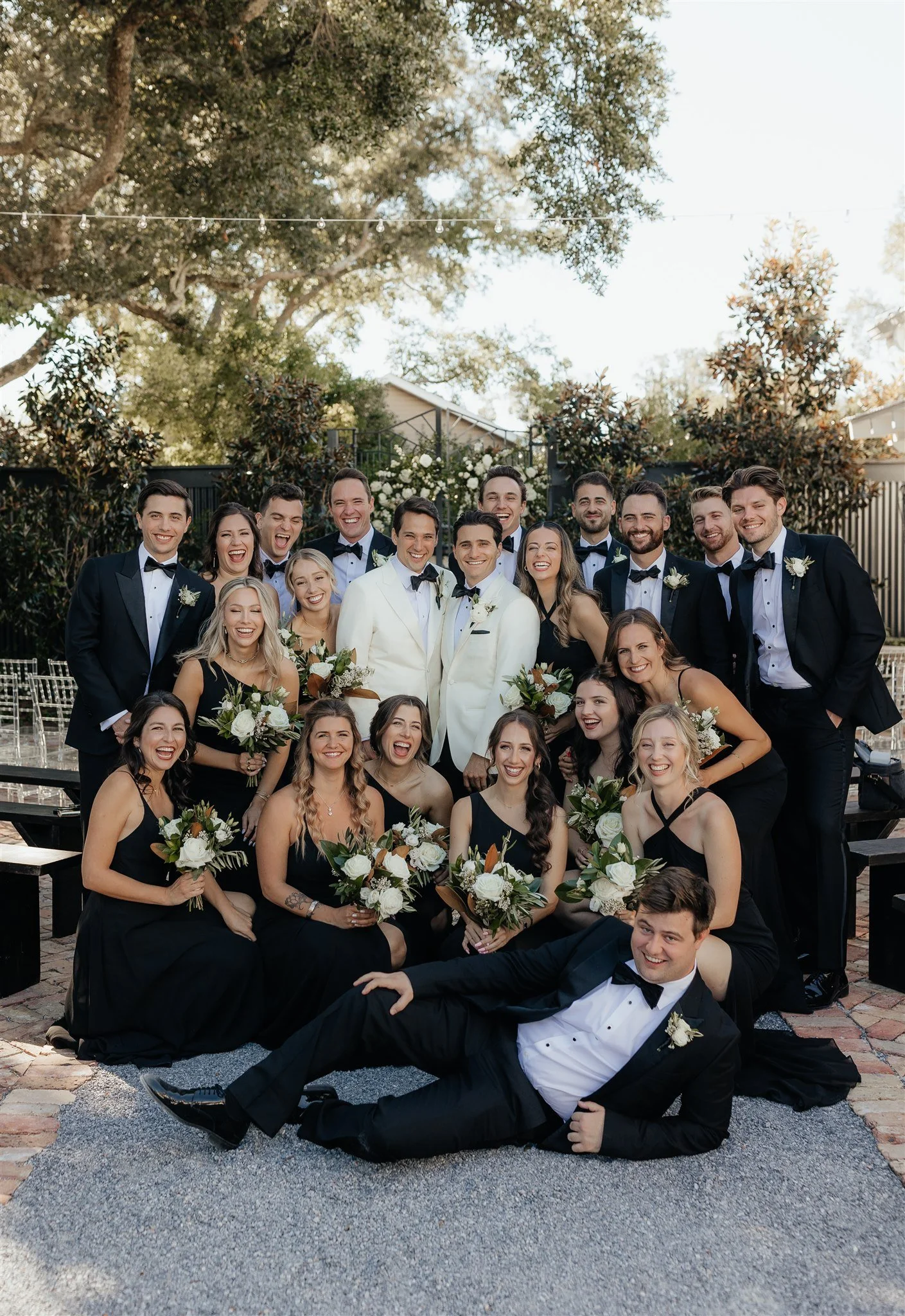 A large Pensacola wedding party outdoors, with men in tuxedos and women in black dresses, holding bouquets of white flowers, smiling and posing together.
