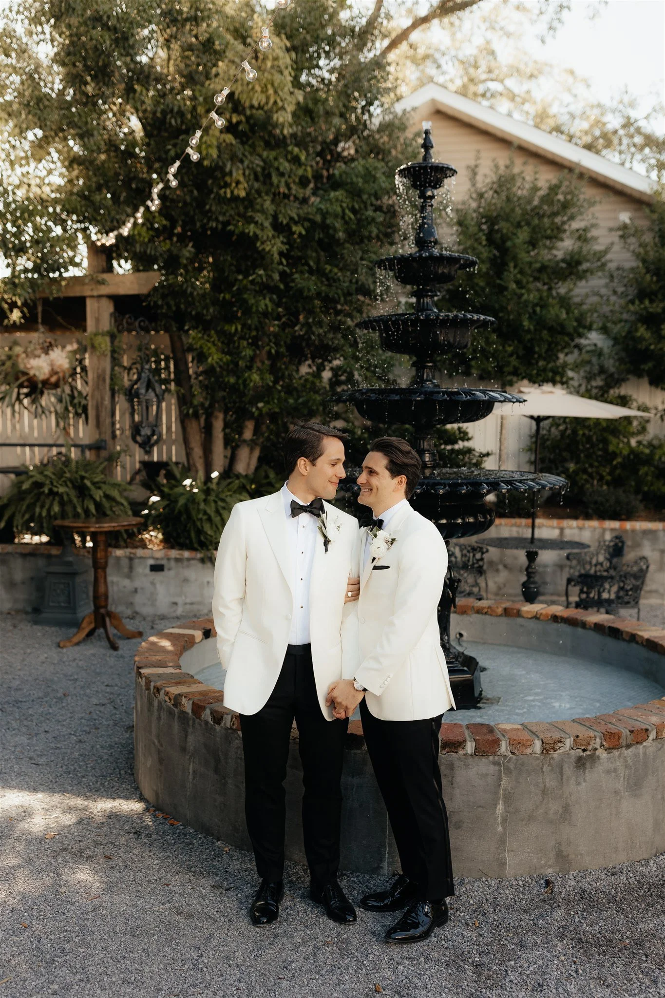 A couple dressed in wedding attire standing in front of a fountain, holding hands, and gazing at each other, outdoors in a garden setting.
