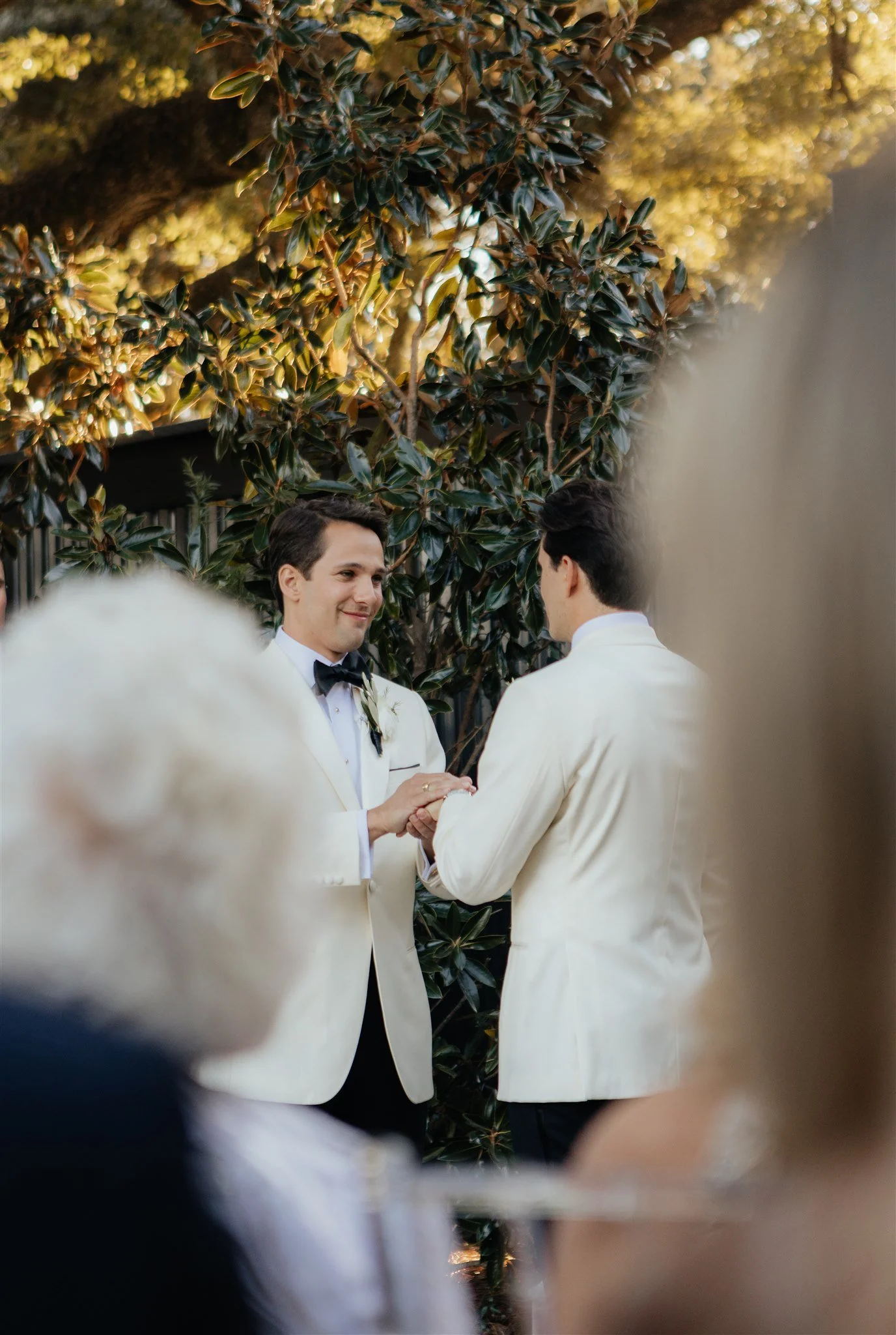 Two men in white tuxedos holding hands during a wedding ceremony outdoors with tall trees and sunlight in the background.