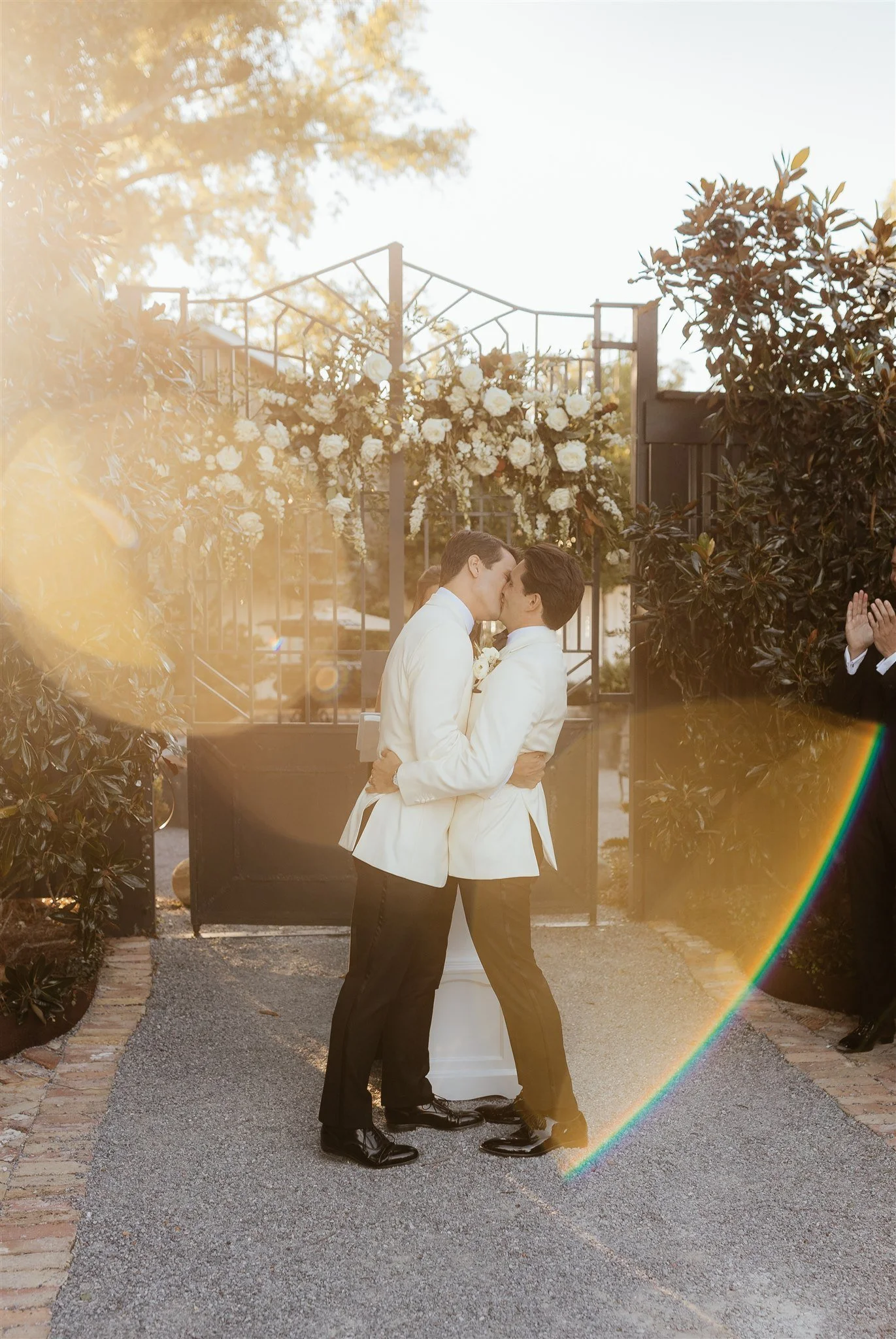 A same-sex couple dressed in white jackets sharing a kiss during their wedding ceremony, with floral decorations and sunlight creating a warm ambiance.