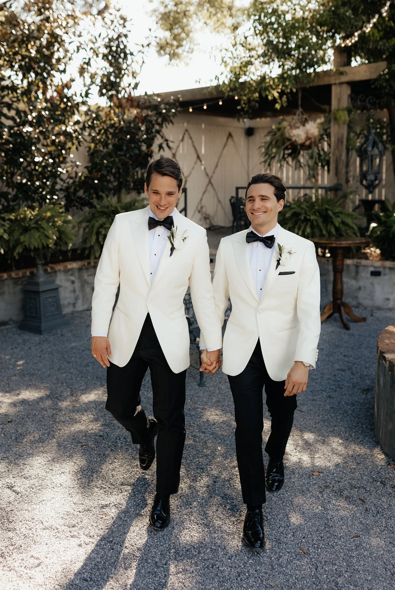 Two men in white tuxedo jackets and black bow ties walking hand in hand outdoors, smiling, with greenery and string lights in the background.