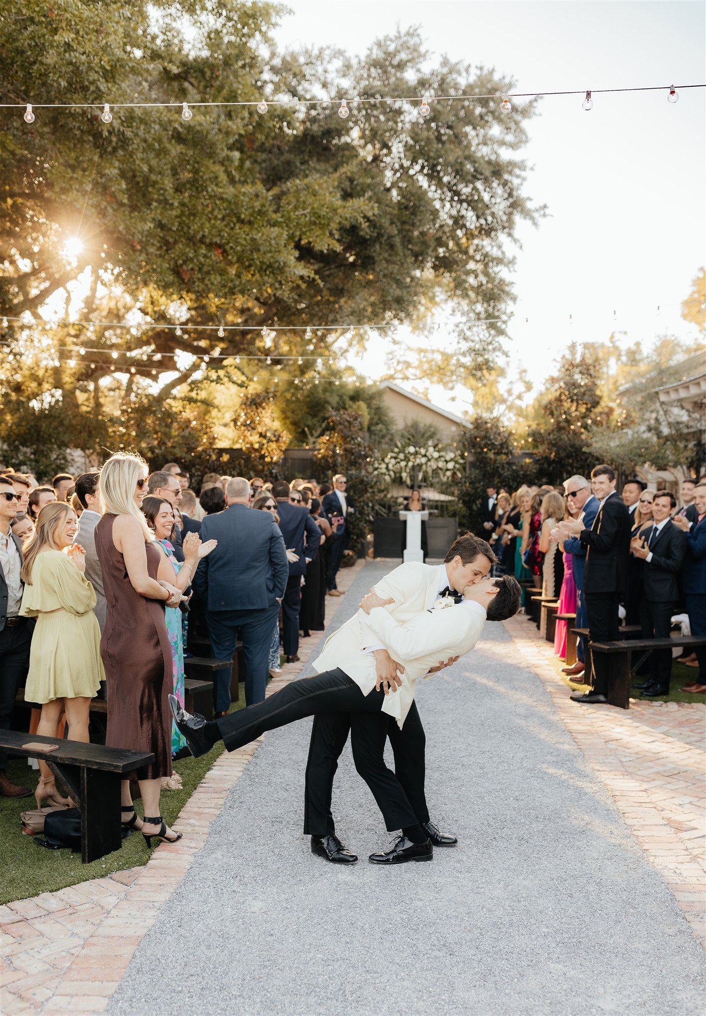 LGBTQ couple kissing at wedding ceremony outdoors with guests clapping and smiling in the background, trees, and string lights.