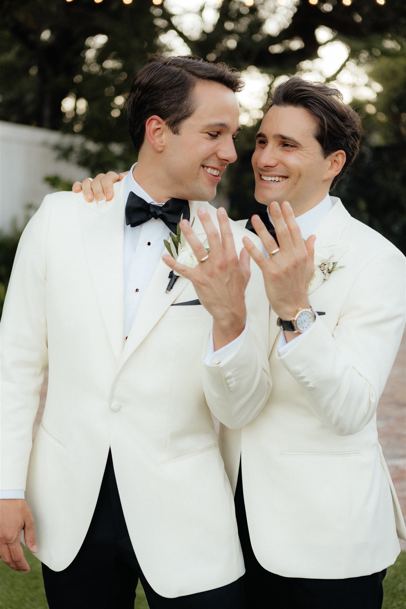 Two men in white tuxedos and black bow ties showcasing wedding rings, smiling and close together outdoors during daytime.