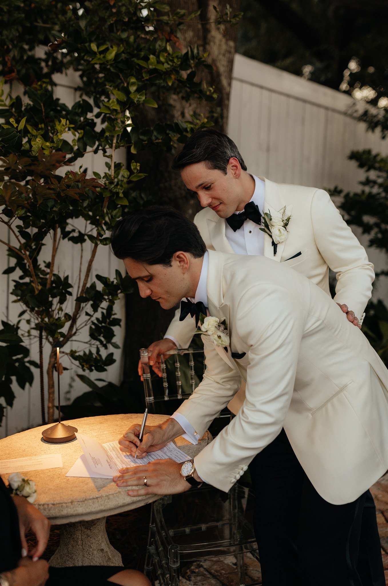Two men in white tuxedos with black bow ties signing a wedding document outdoors, one leaning over the table and the other standing behind him, with a white fence, trees, and greenery in the background.