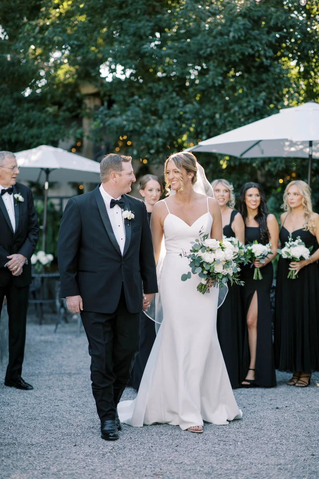 A bride and groom walking hand in hand at their outdoor wedding ceremony, smiling at each other, surrounded by bridesmaids and groomsmen, with umbrellas and trees in the background.