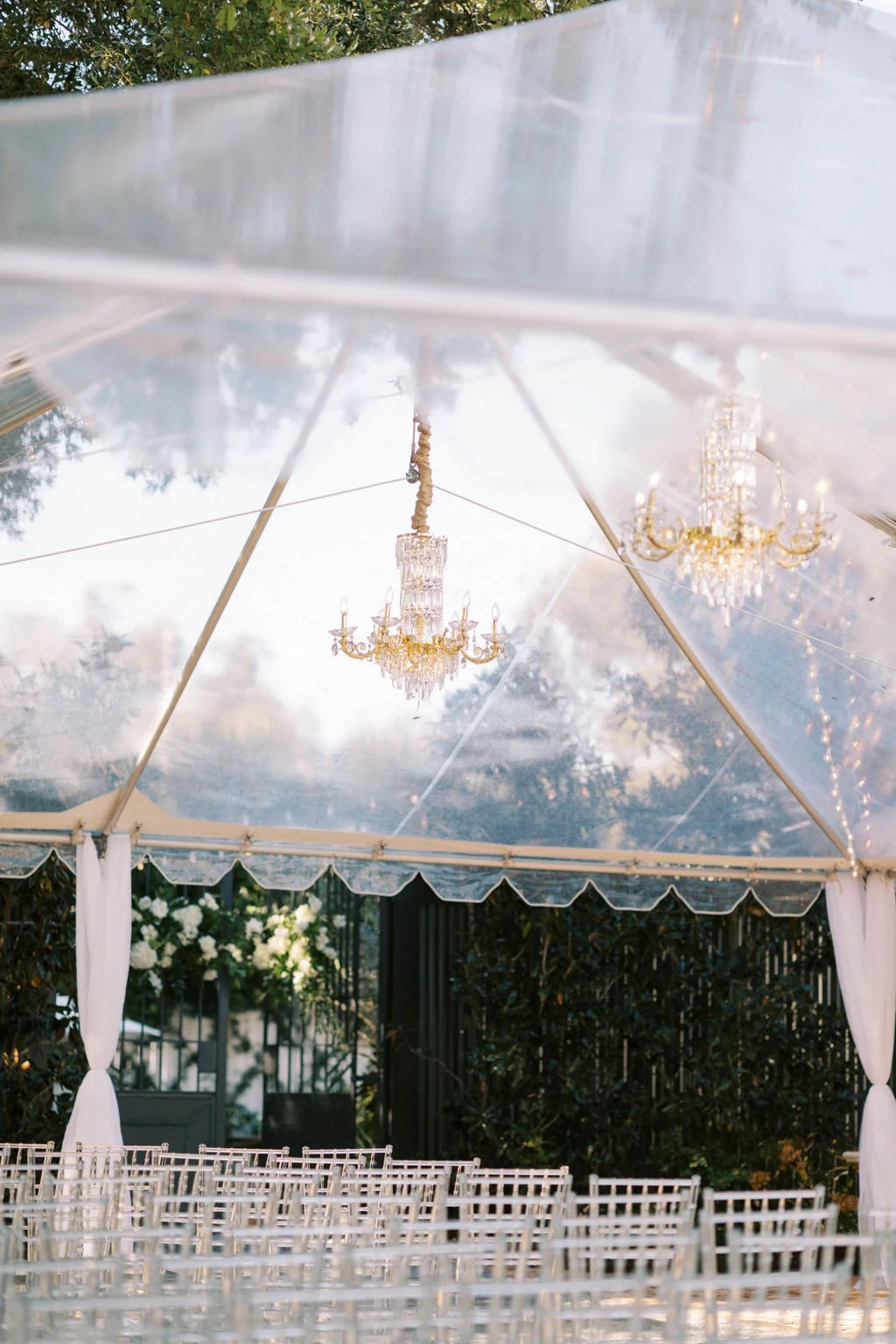 Elegant outdoor wedding setup with a clear canopy, white chairs, and chandeliers hanging from the ceiling, surrounded by greenery.