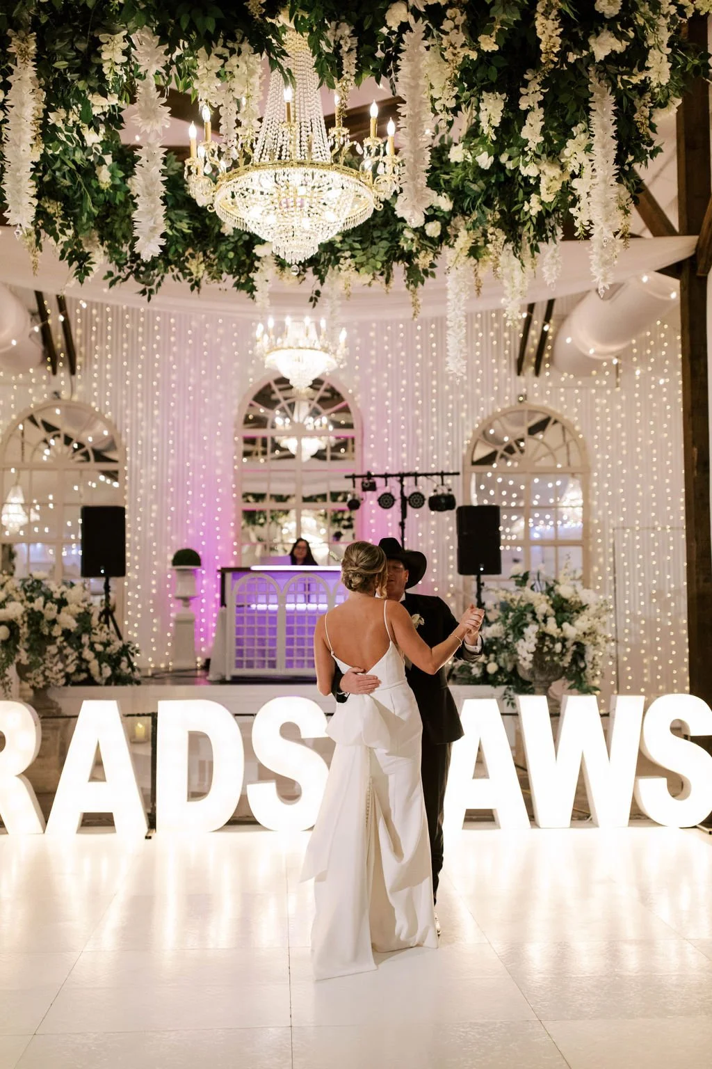 A Pensacola bride and groom dancing at their wedding reception with large illuminated letters reading 'GIRADS AWS' in front of them, surrounded by floral decorations and chandeliers.
