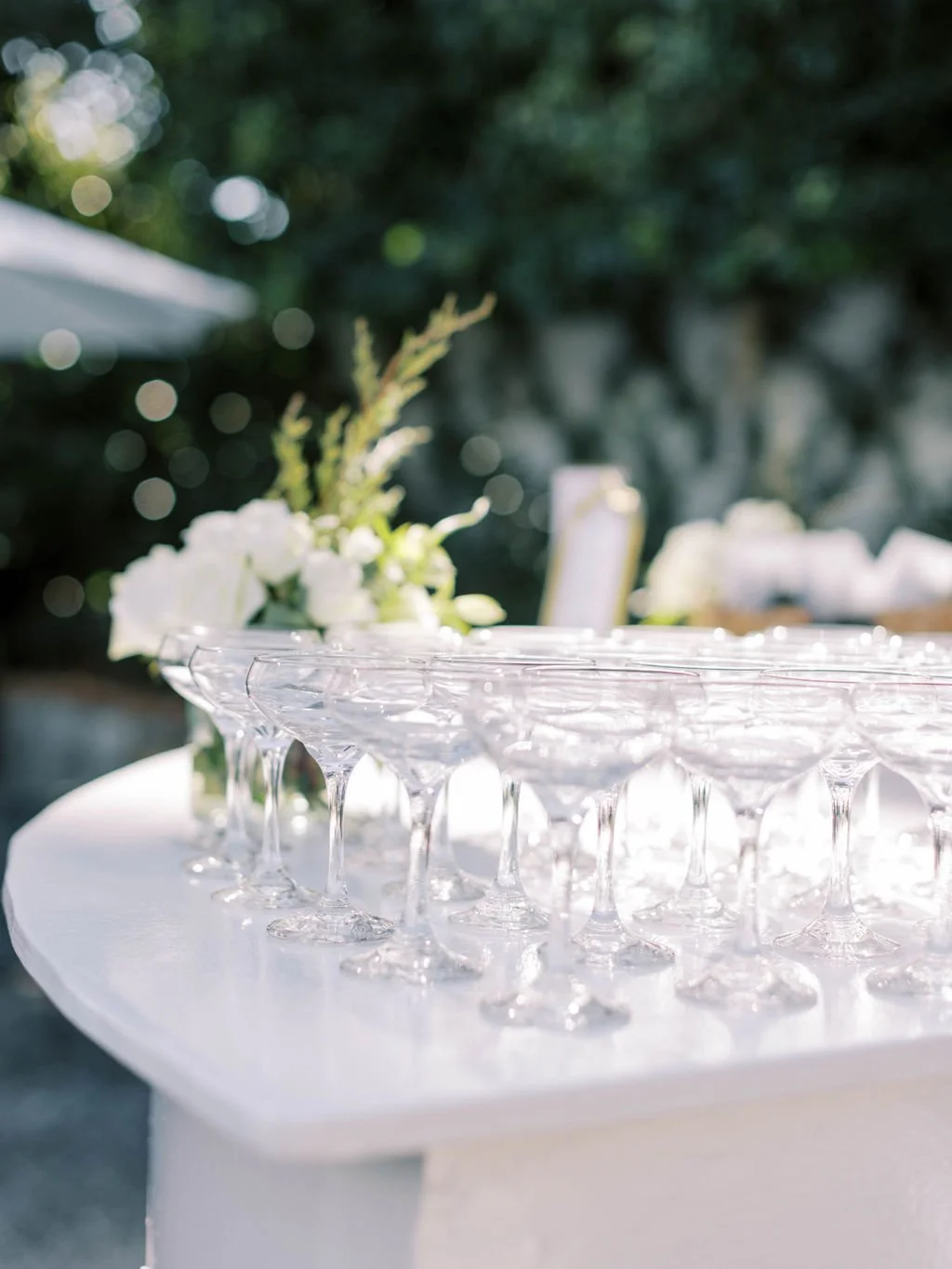 Multiple empty champagne glasses on a white tablecloth with a blurred floral arrangement in the background at an outdoor event.