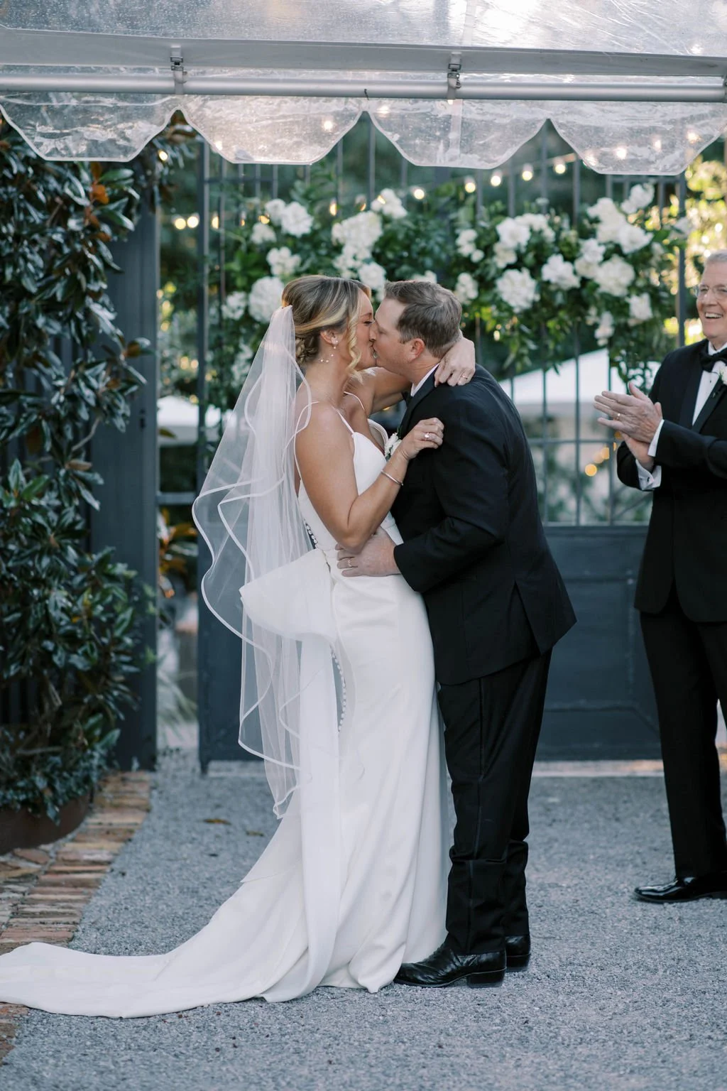 A Pensacola bride and groom share a kiss during their wedding ceremony, with a clapping man in a tuxedo on the right and floral decorations in the background.
