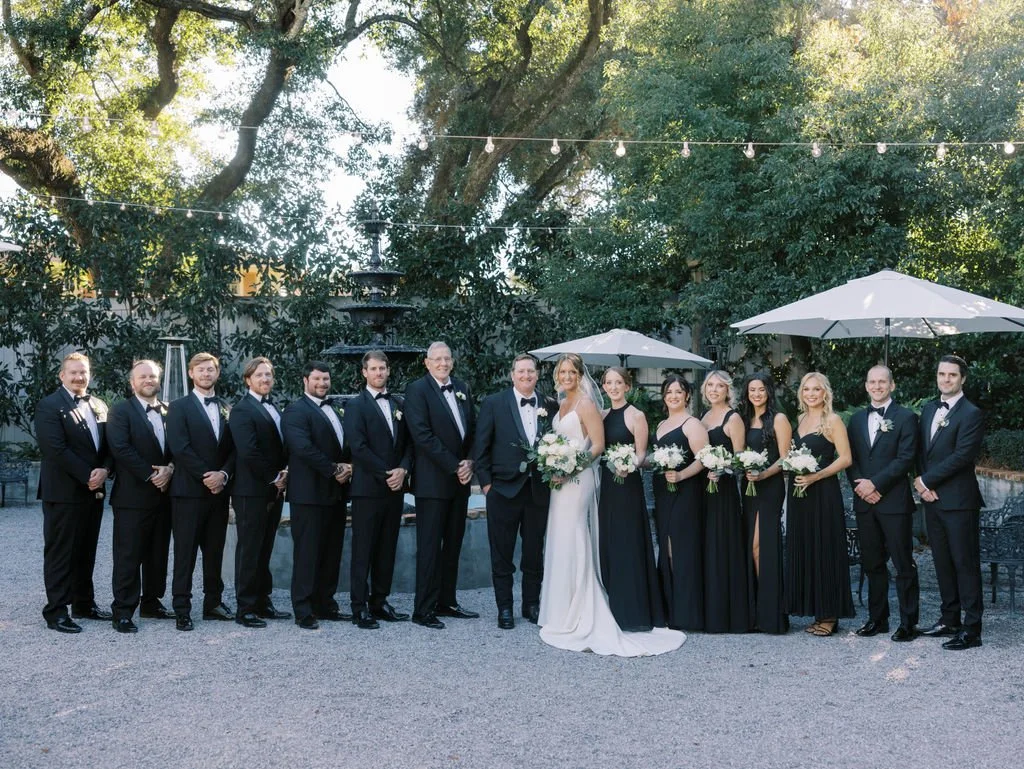 A Pensacola wedding party with 13 people standing outdoors, with trees, string lights, and umbrellas in the background. The bride and groom are in the center, surrounded by bridesmaids and groomsmen dressed in formal attire.