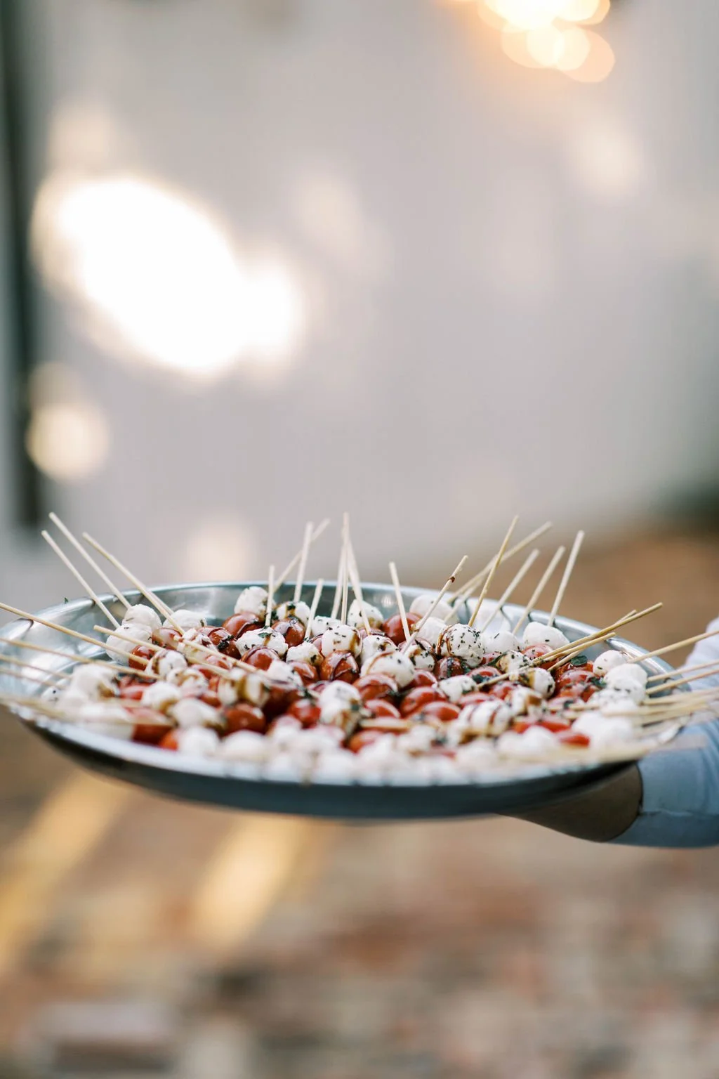 A person holding a silver tray of assorted skewered appetizers, including cherry tomatoes and mozzarella balls, at a blurred outdoor setting with warm lighting.