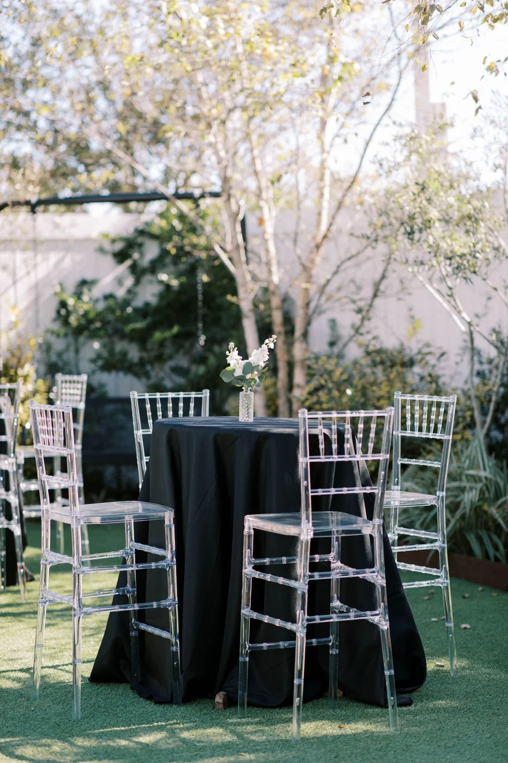 Outdoor table setup with a black tablecloth, clear ghost chairs, and a vase with white flowers in a garden with trees and plants in a Pensacola Wedding Venue.