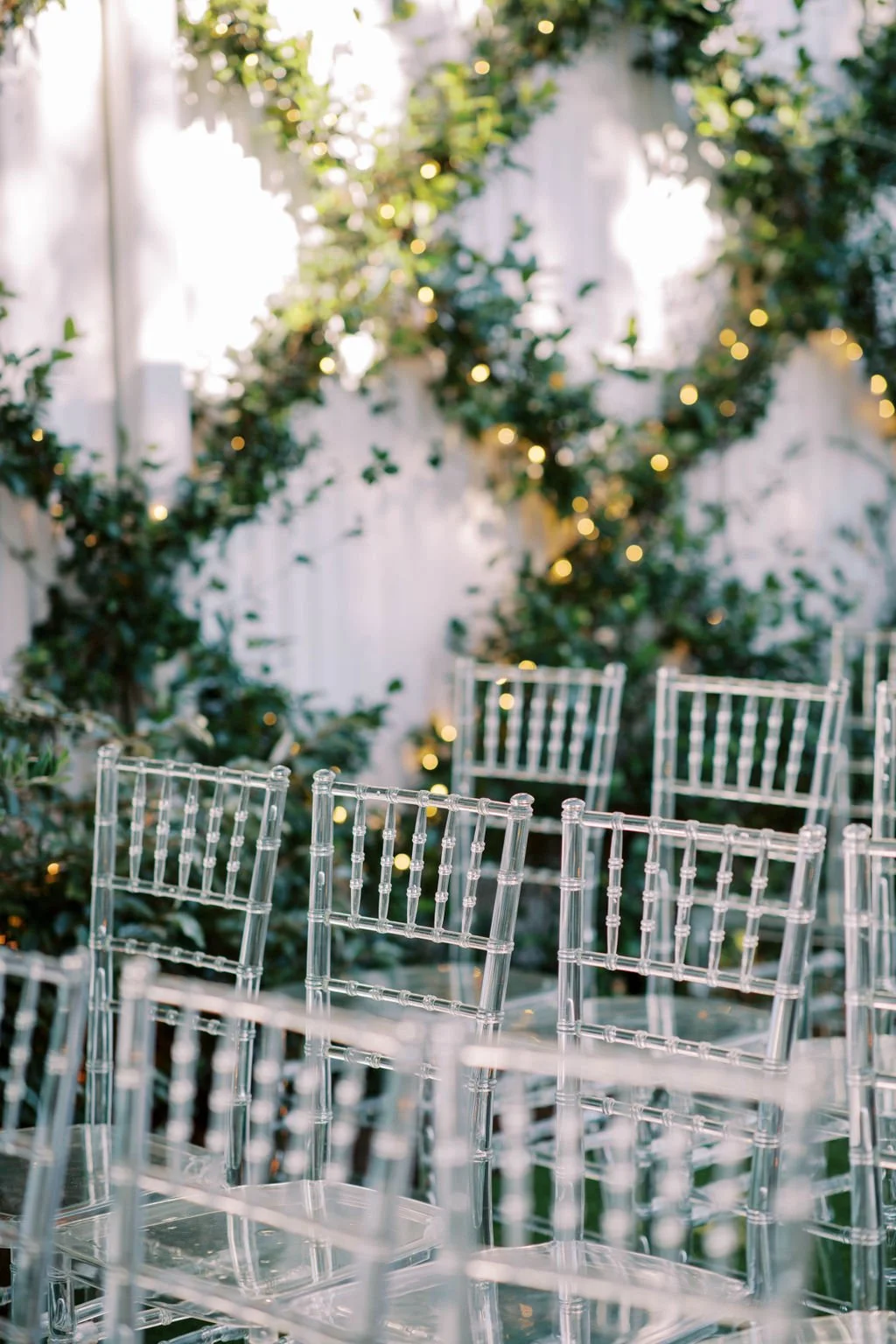 Clear acrylic chairs arranged outdoors with string lights and greenery in the background, likely set for an event.