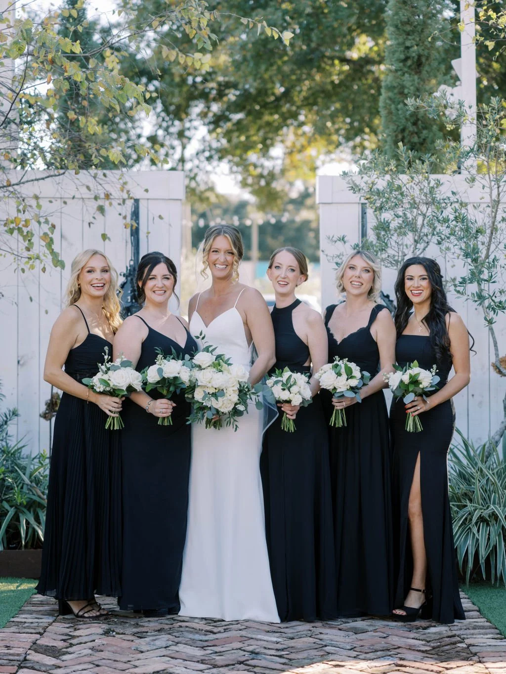 A bride in a white wedding dress with a bouquet of white flowers, surrounded by five bridesmaids in long black dresses holding smaller bouquets, standing outdoors on a cobblestone path with white fences and greenery.