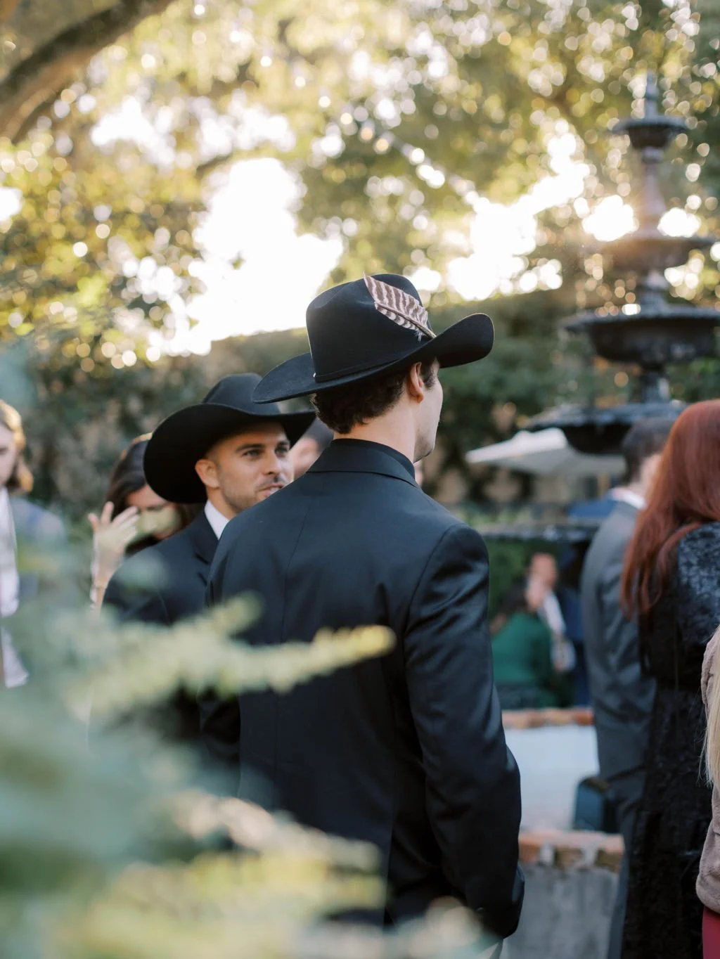 People dressed in formal attire with wide-brimmed hats, standing outdoors near a fountain with trees and sunlight in the background.