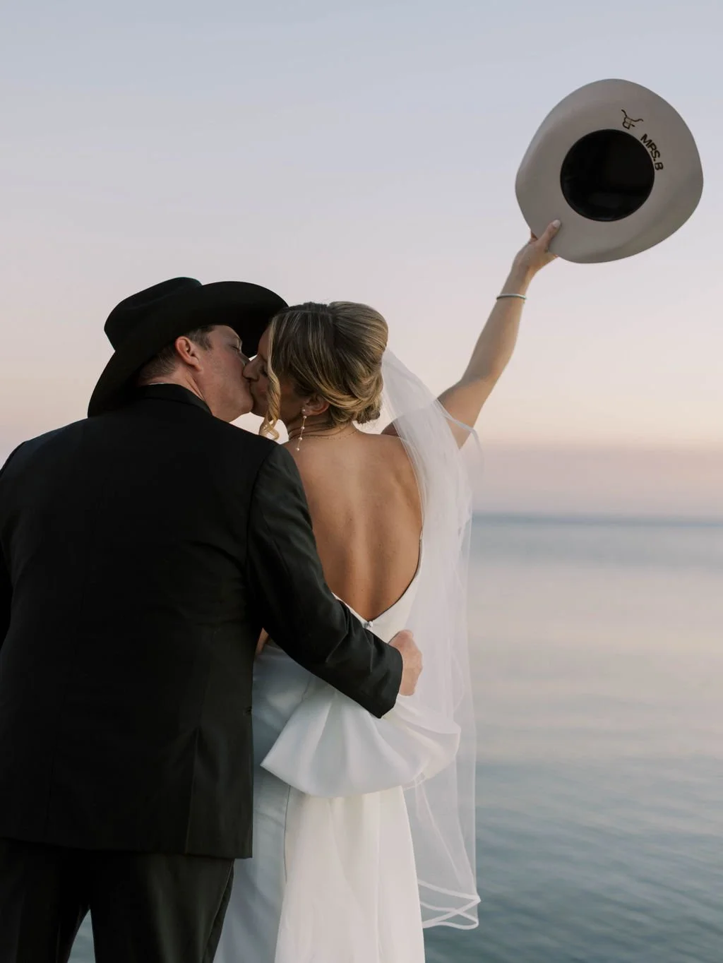 A couple in wedding attire sharing a kiss during sunset by the water, with the bride holding a cowboy hat in the air.