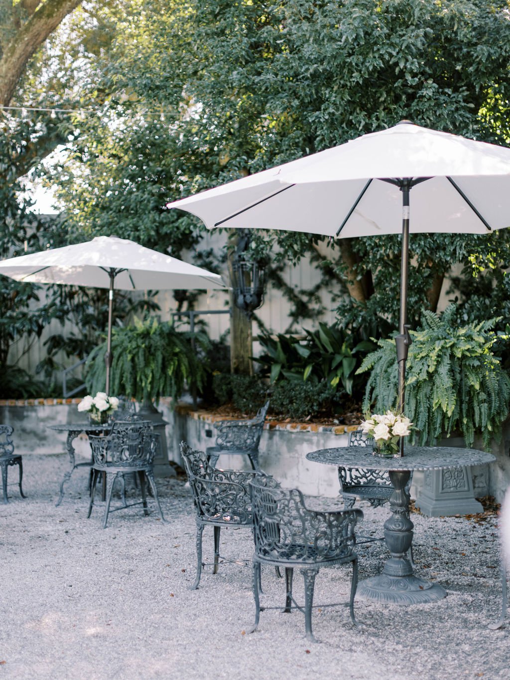 Outdoor patio with ornate metal tables and chairs, large white umbrellas, and potted flowers surrounded by lush green trees and plants in a Pensacola Wedding Venue.