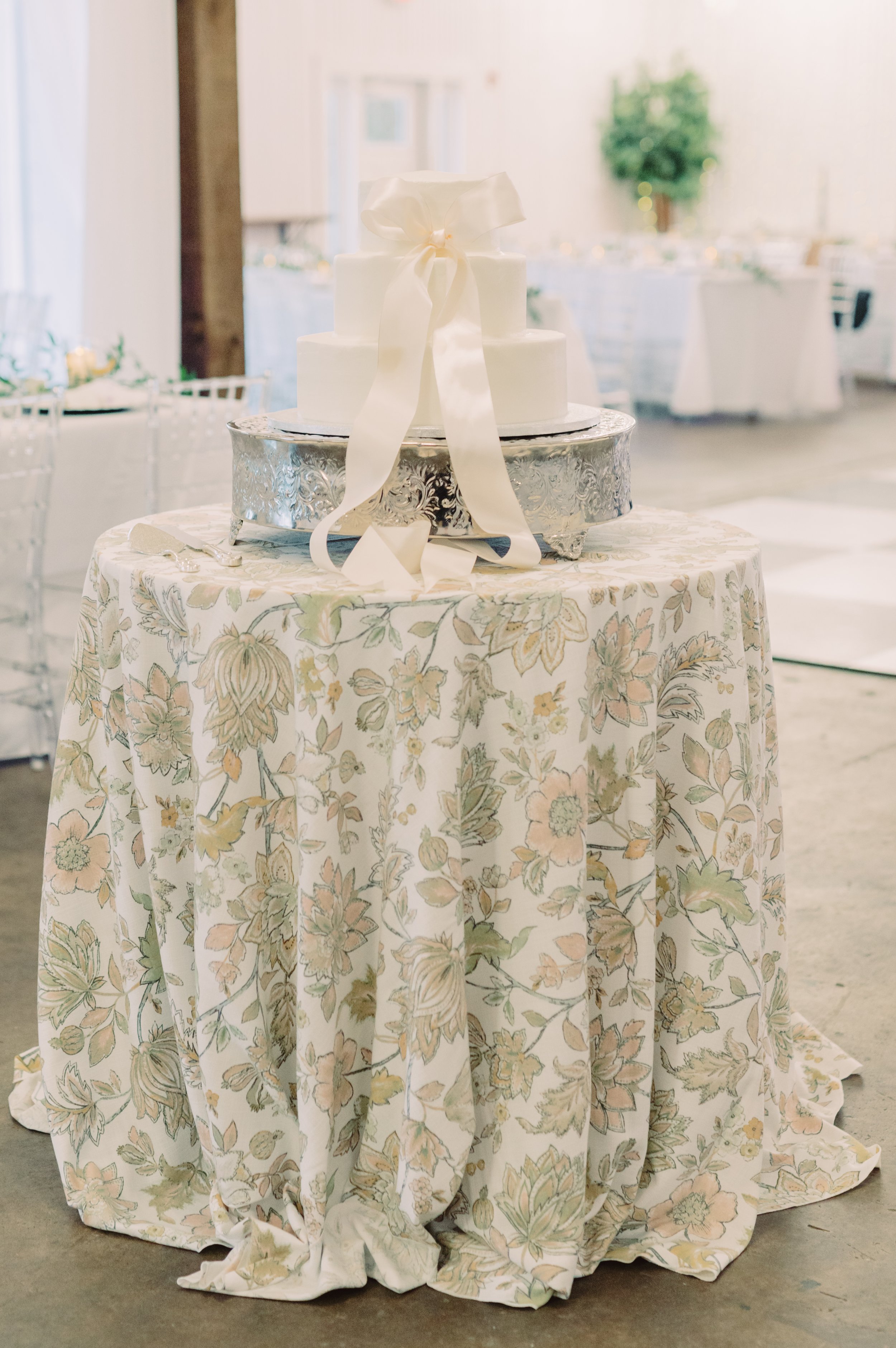 A three-tiered white wedding cake decorated with a large satin bow on top, displayed on a decorative silver cake stand, placed on a round table covered with a floral tablecloth in a decorated banquet hall.
