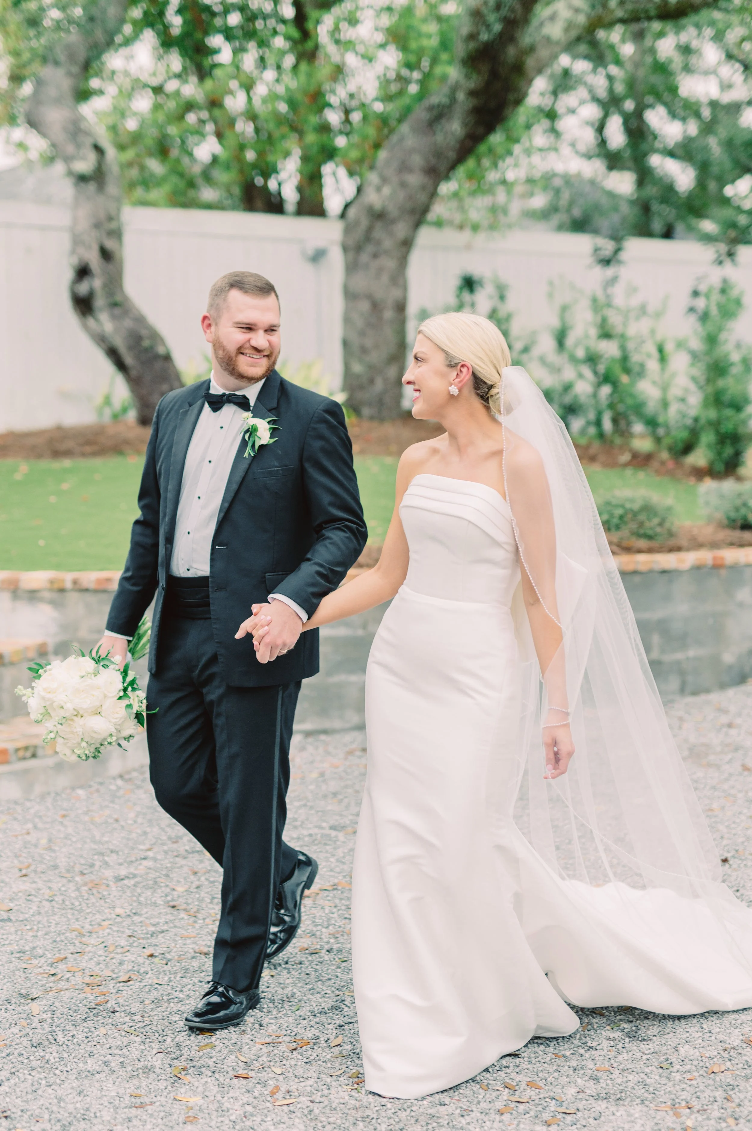 A newlywed couple walking outdoors holding hands, smiling at each other. The groom wears a black tuxedo with a bow tie, and the bride wears a strapless white wedding gown with a veil. The bride holds a bouquet of white flowers, and they are surrounde