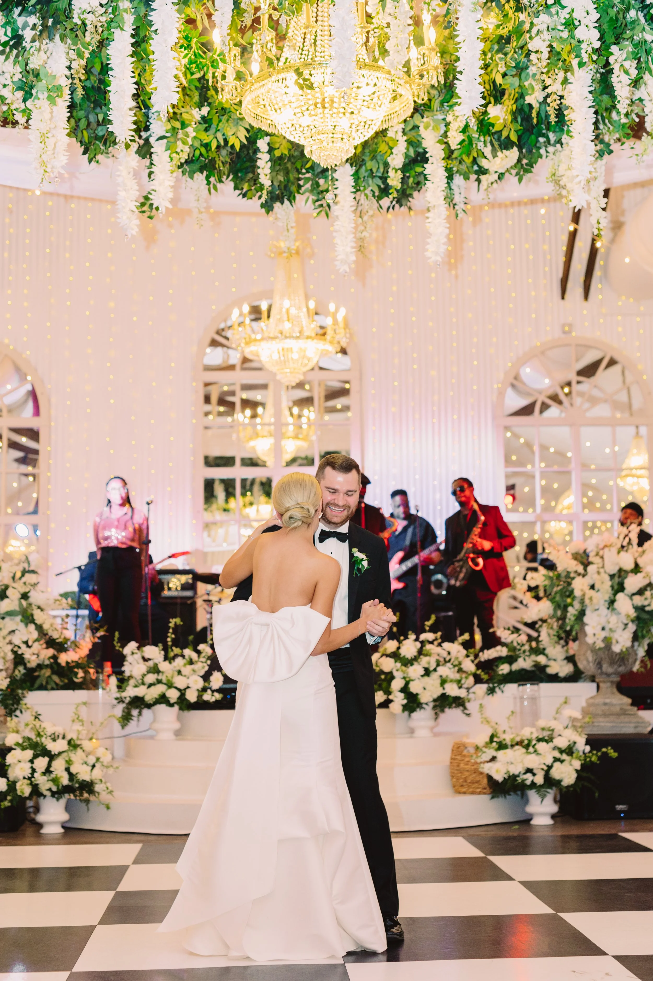 A newlywed couple dancing at their wedding reception, surrounded by floral arrangements and illuminated by chandeliers. The bride in a white gown and the groom in a black tuxedo are smiling, with a live band performing in the background.