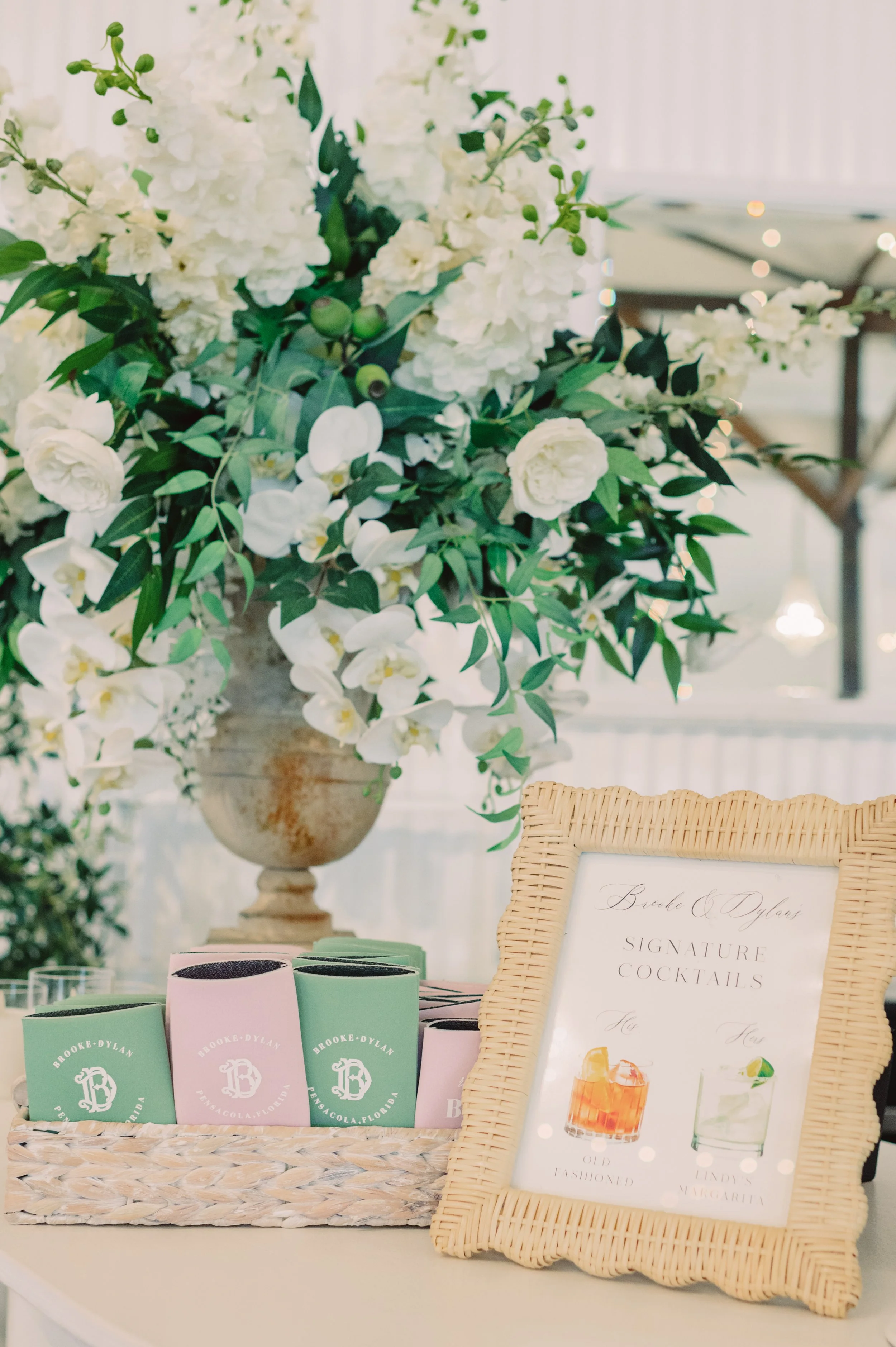 A floral arrangement with white flowers and greenery, a framed sign for signature cocktails, and a basket of pink and green drink napkins.