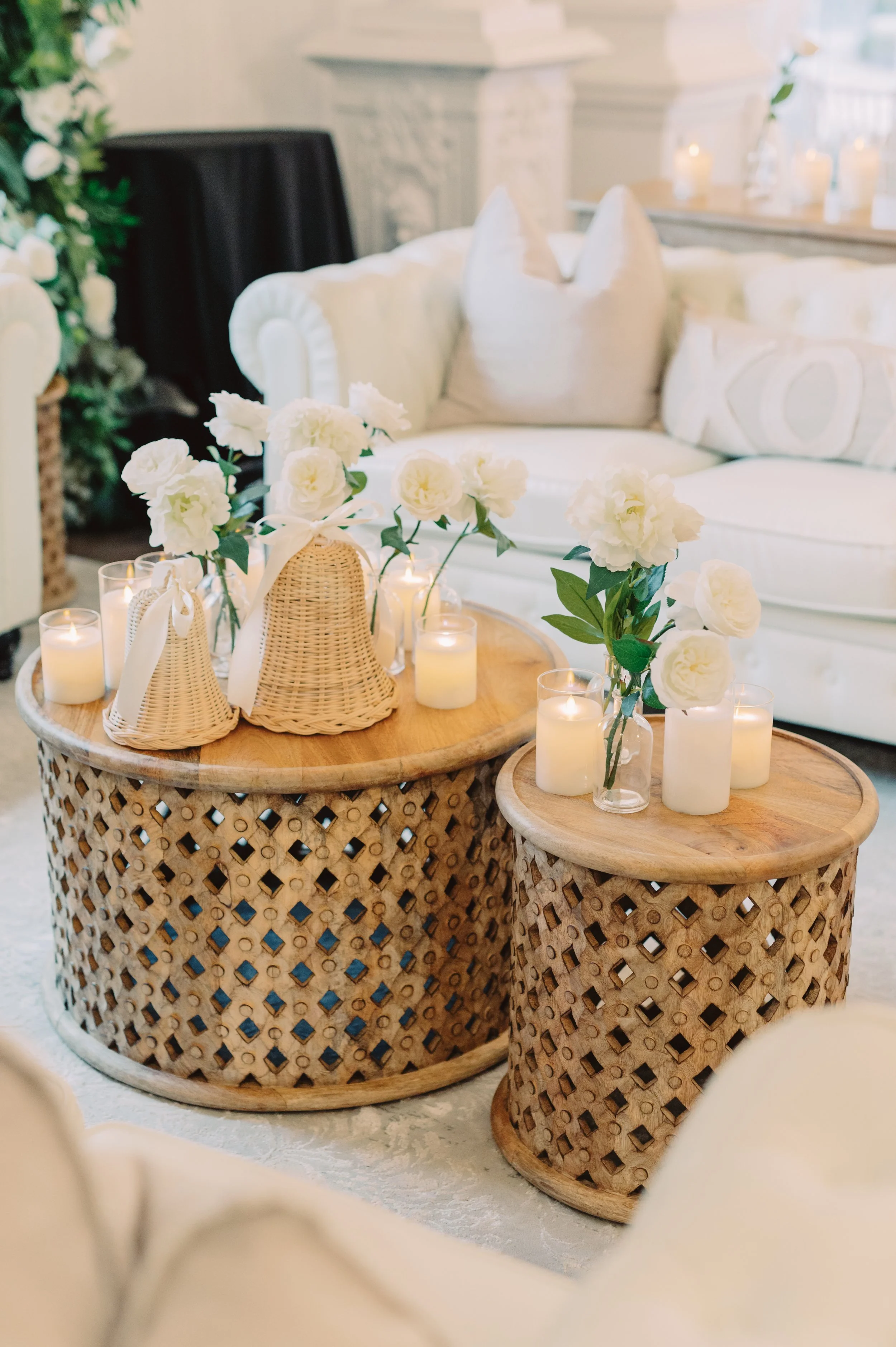 Decorative Pensacola Wedding Venue with two wicker coffee tables topped with white flowers in glass vases and lit candles, white sofa with pillows, and candles on a mantel in the background.