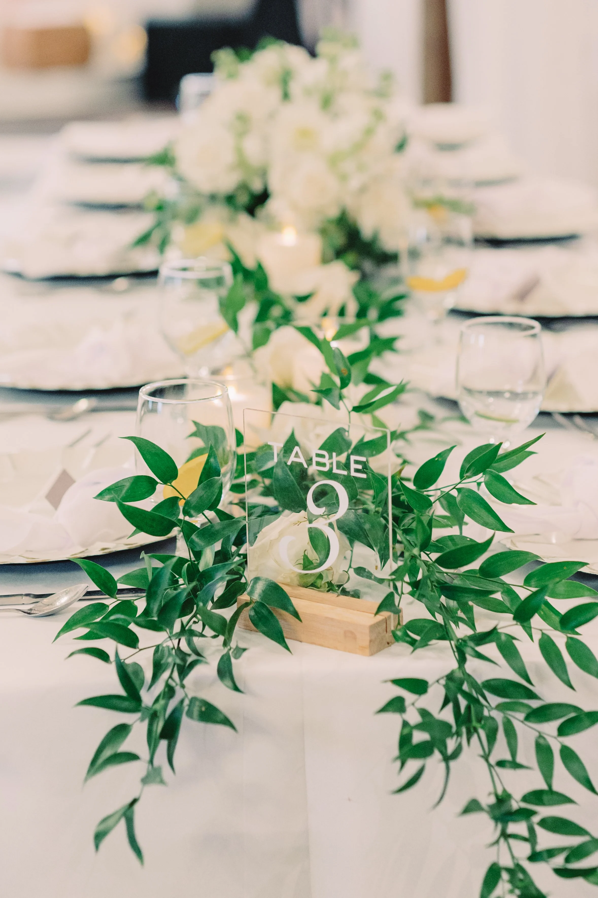 Wedding reception table centerpiece with greenery and a table number 3 sign.