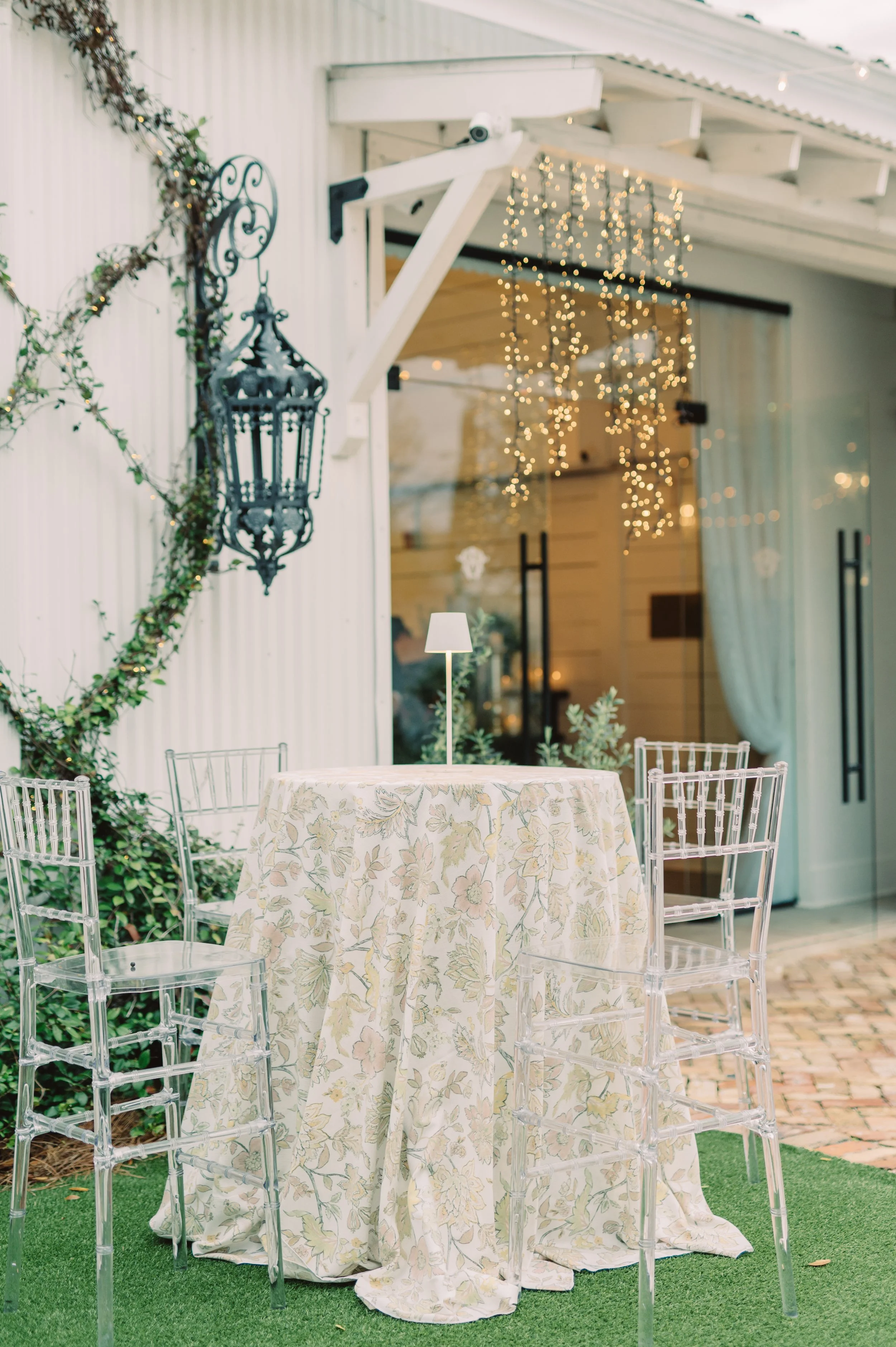 Outdoor table with a floral tablecloth and a small lamp, surrounded by clear acrylic chairs, with string lights and a lantern on the wall in the background.